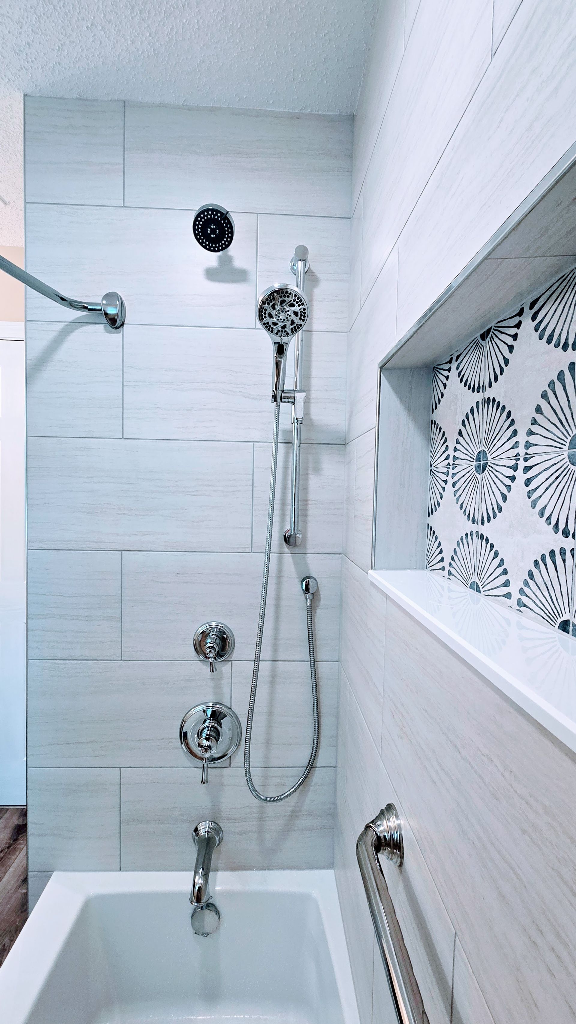 Bathroom shower with white tiles, hand shower, built-in shelf with patterned tile, and grab bar.