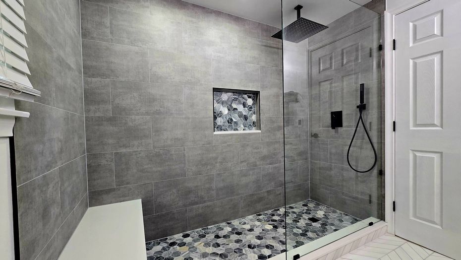 Gray-tiled shower in West Hartford, CT with pebble floor, glass door, and built-in shelf. Black showerhead and handheld wand. White door to the right.