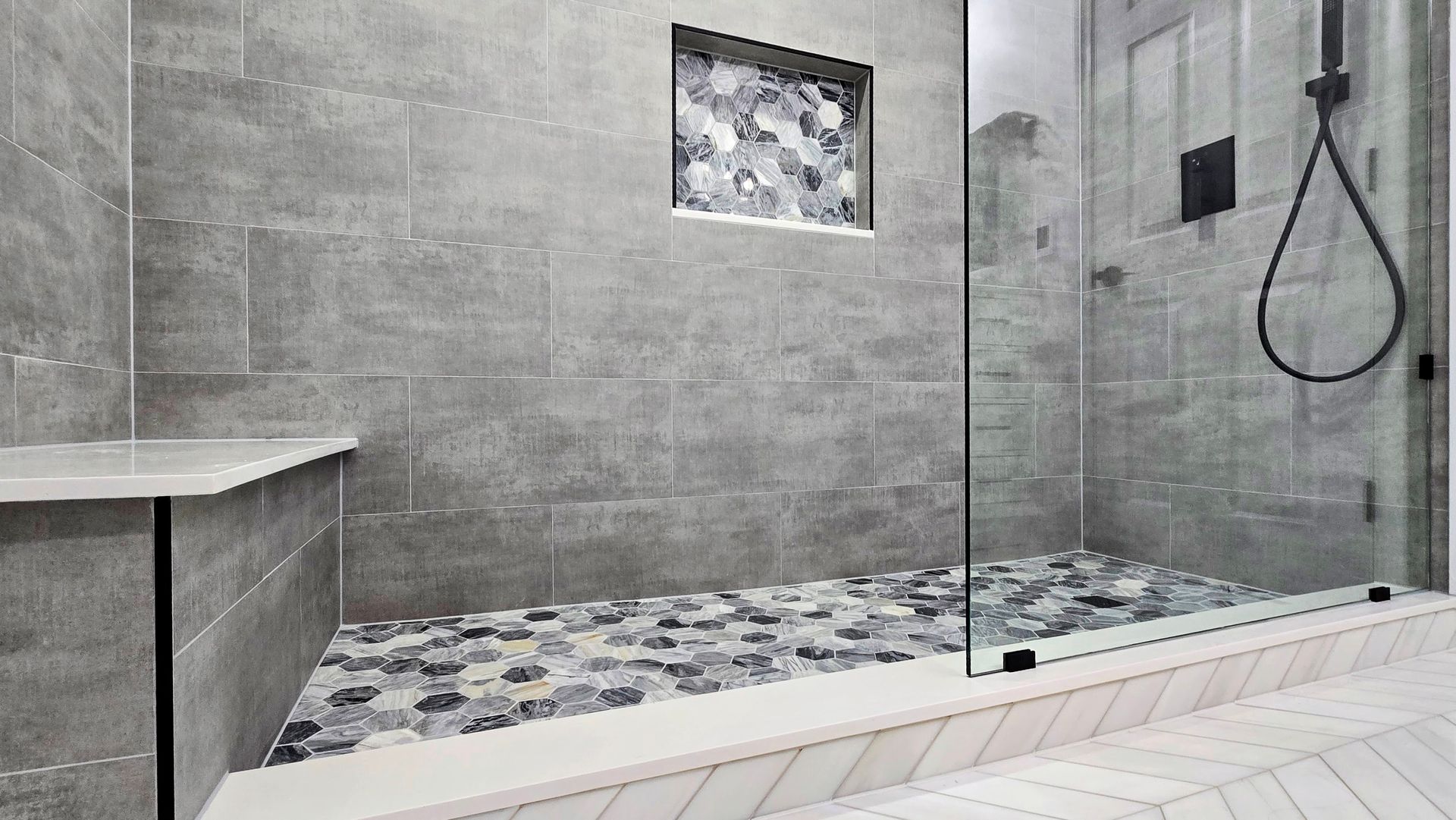 Gray tiled bathroom with glass shower, pebbled floor, built-in bench, and decorative niche.