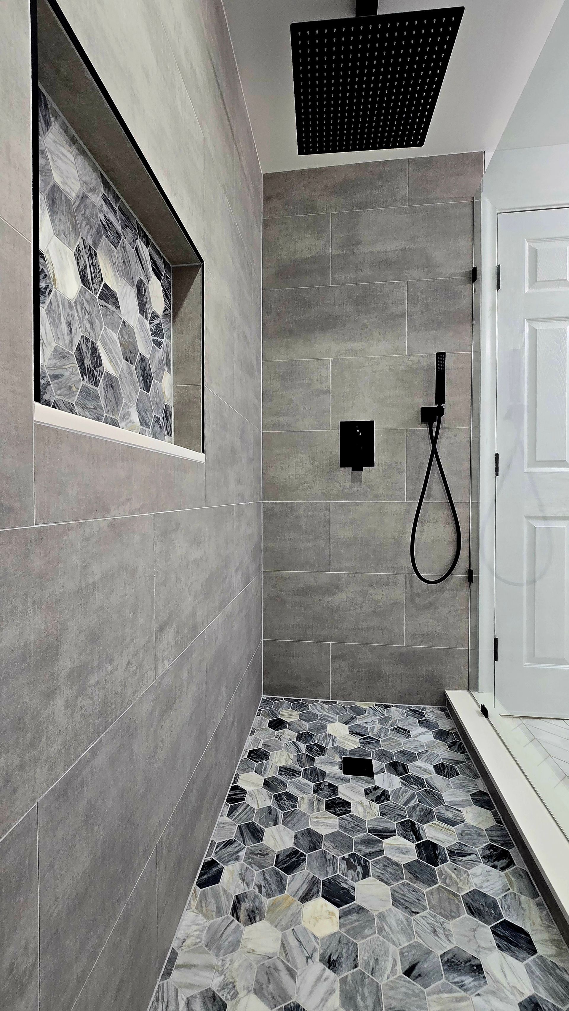 A modern walk-in shower in West Hartford, CT with large grey wall tiles, a mosaic hexagon tile floor, and black fixtures, including a niche.