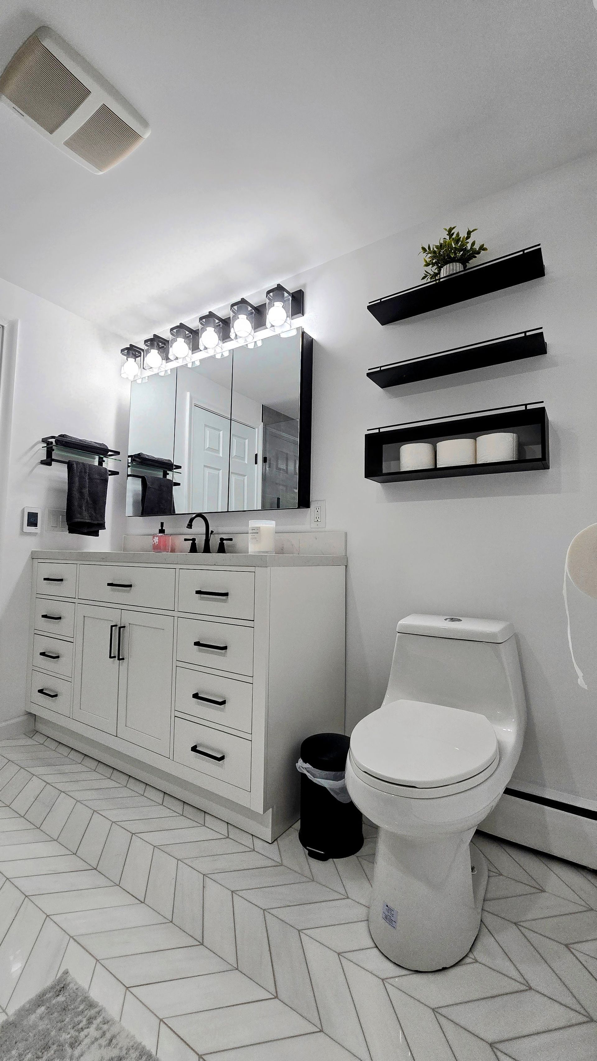 A bright bathroom remodel in West Hartford, CT features a white vanity, a large mirror, a toilet, and three black wall shelves on white herringbone tile.