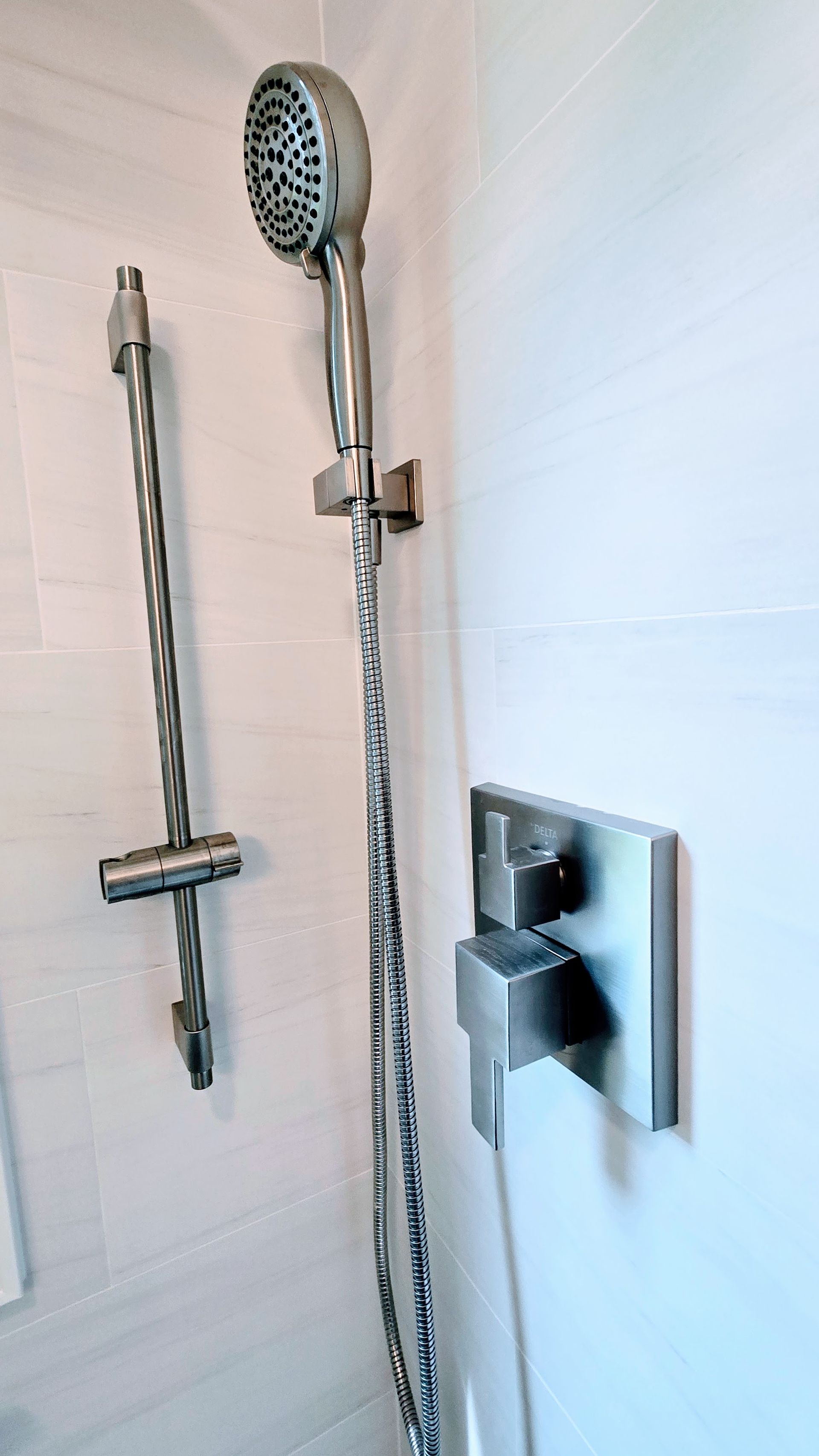 Showerhead and valve on a white-tiled wall. A handheld showerhead hangs, and a temperature/flow control lever is on the right.