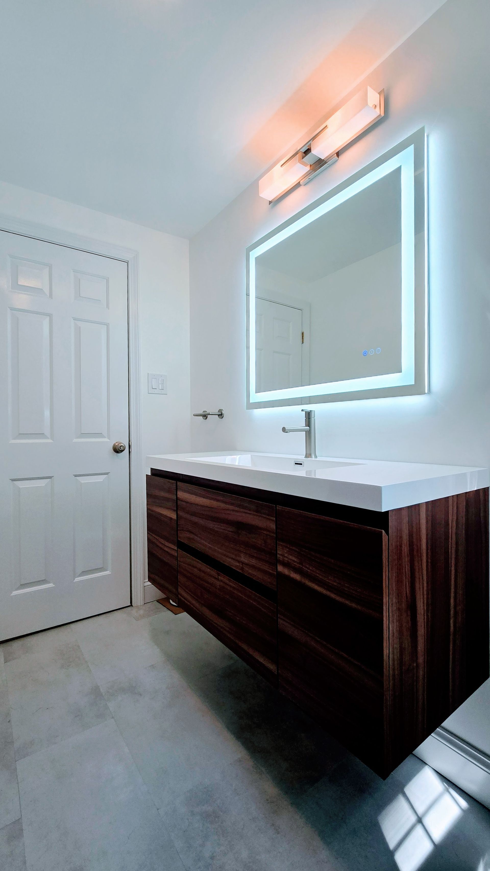 Modern bathroom in West Simsbury with a floating wooden vanity, illuminated mirror, and light gray tile flooring. A white door is on the left.