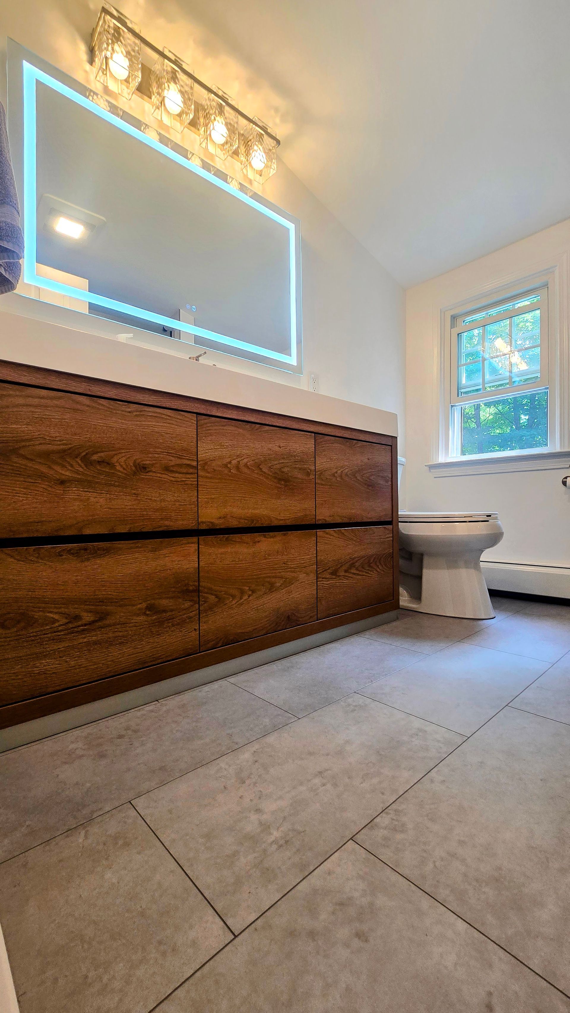 West Simsbury Bathroom with wood vanity, large mirror, gray tile floor, and toilet near a window.