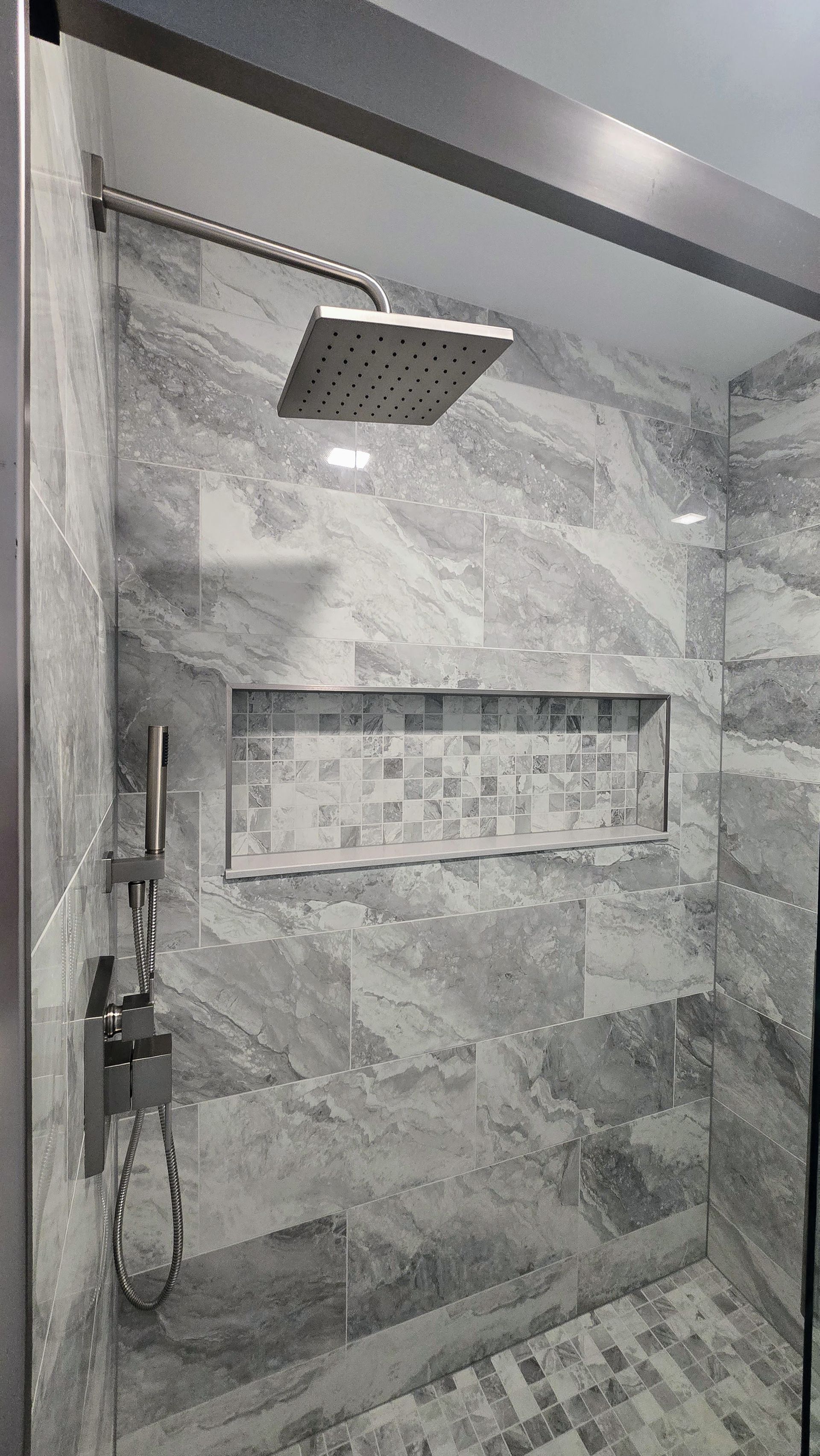 Gray tiled shower with a niche and showerhead. Silver fixtures. Glass door.