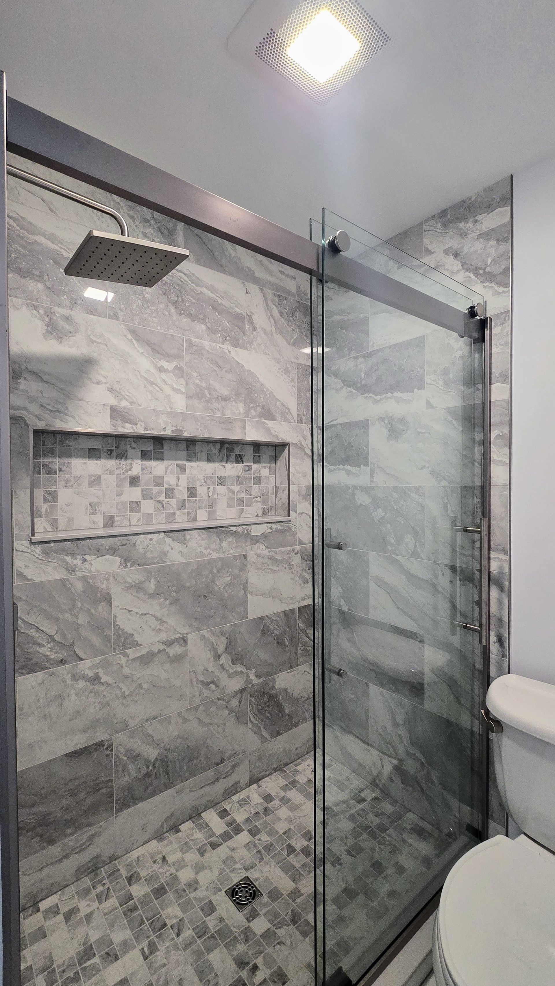 Gray-tiled shower with a sliding glass door and recessed shelf, square rain showerhead. White toilet beside it in Plainville, CT