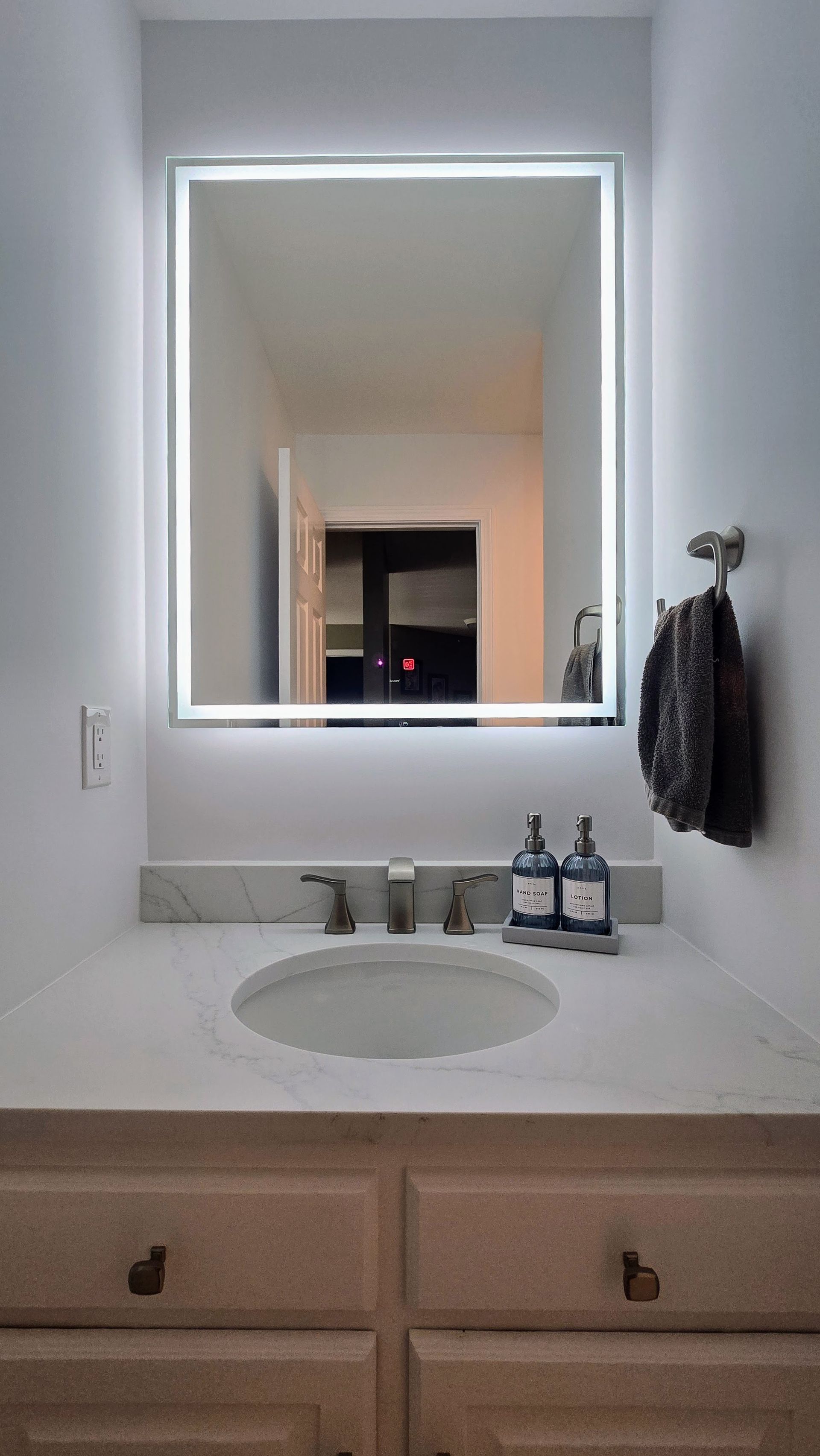 Modern bathroom with lit-up square mirror above a white countertop, sink, and cabinets; a towel rack on the right in Plainville, CT