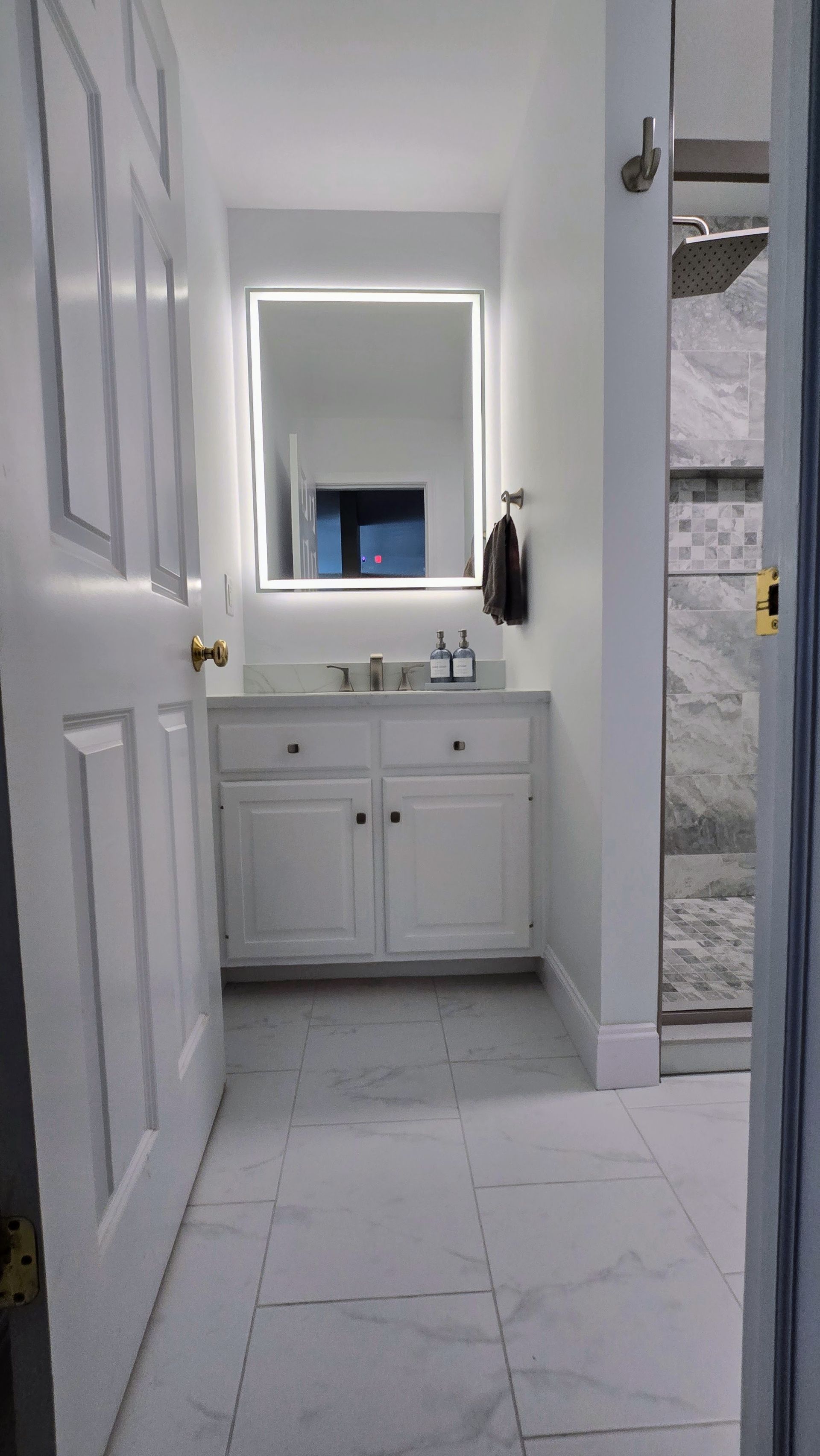 White bathroom with illuminated mirror, white vanity, and marble floor, viewed from doorway.