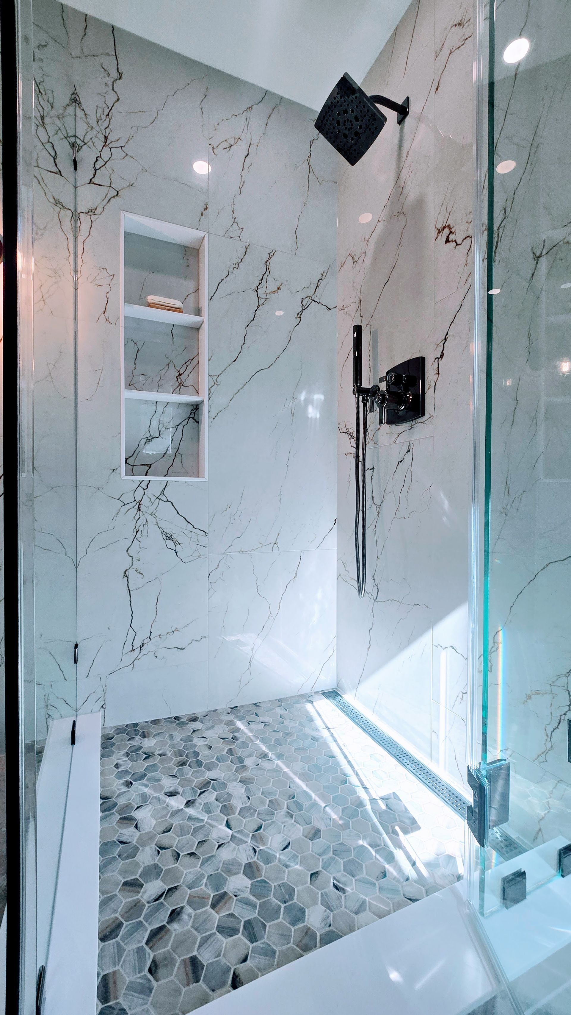 Modern shower remodel in Avon, CT with marble-look wall tiles, pebble floor, glass door, and black shower fixtures.