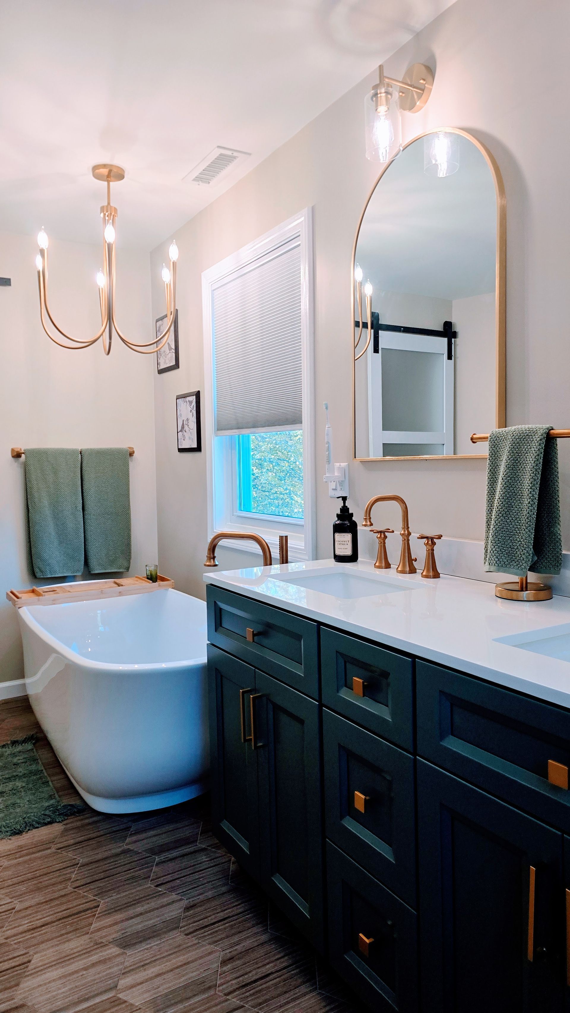 Elegant bathroom renovation in Wolcott, CT with a dark green vanity, white tub, gold accents, and a large arched mirror.