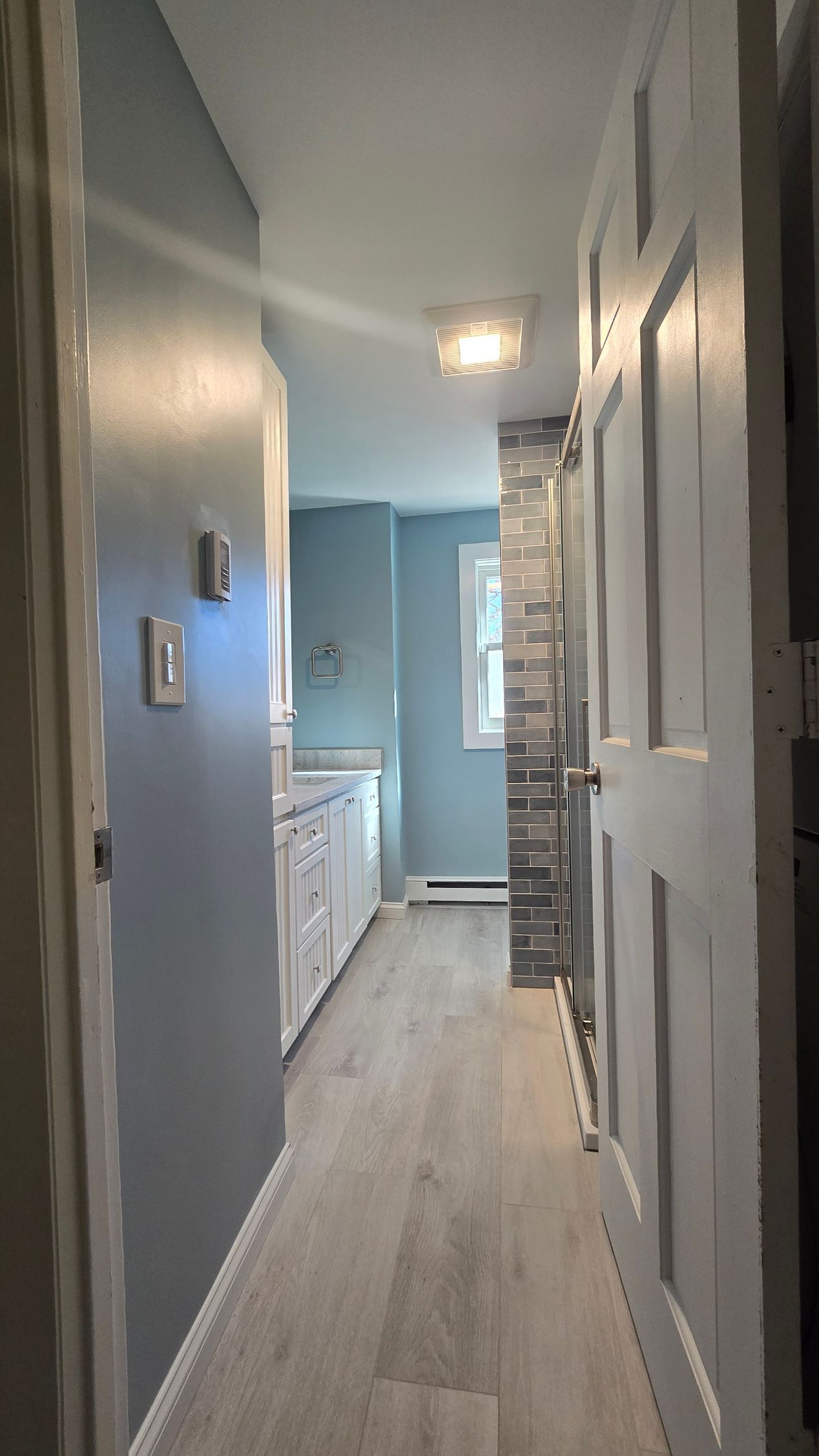 Small bathroom remodel in Ashford, CT with narrow hallway with white doors, light gray floor, and pale blue walls leading to a window.