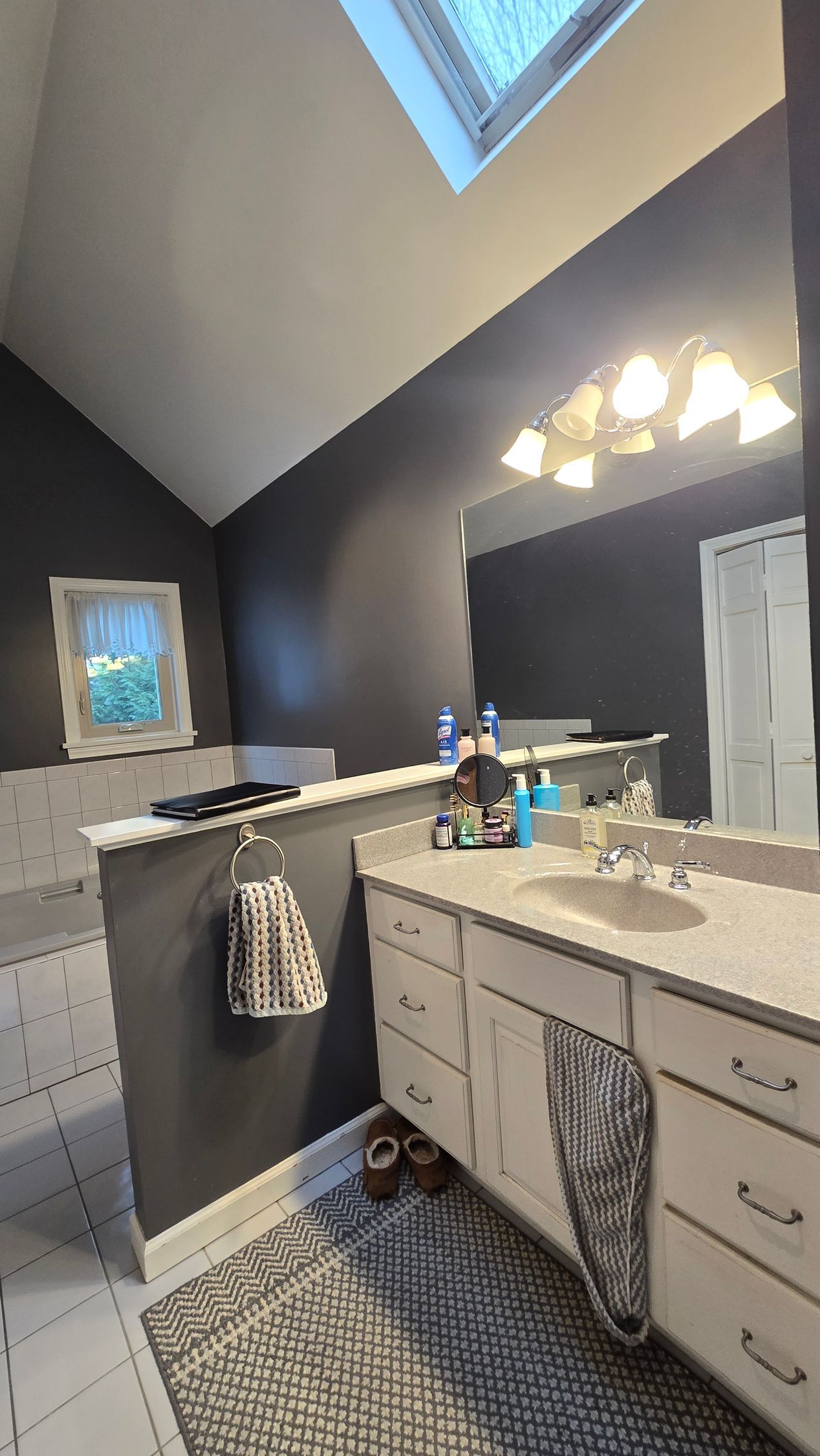 Bathroom vanity with double sinks, dark walls, and a skylight above a bright mirror and light fixtures