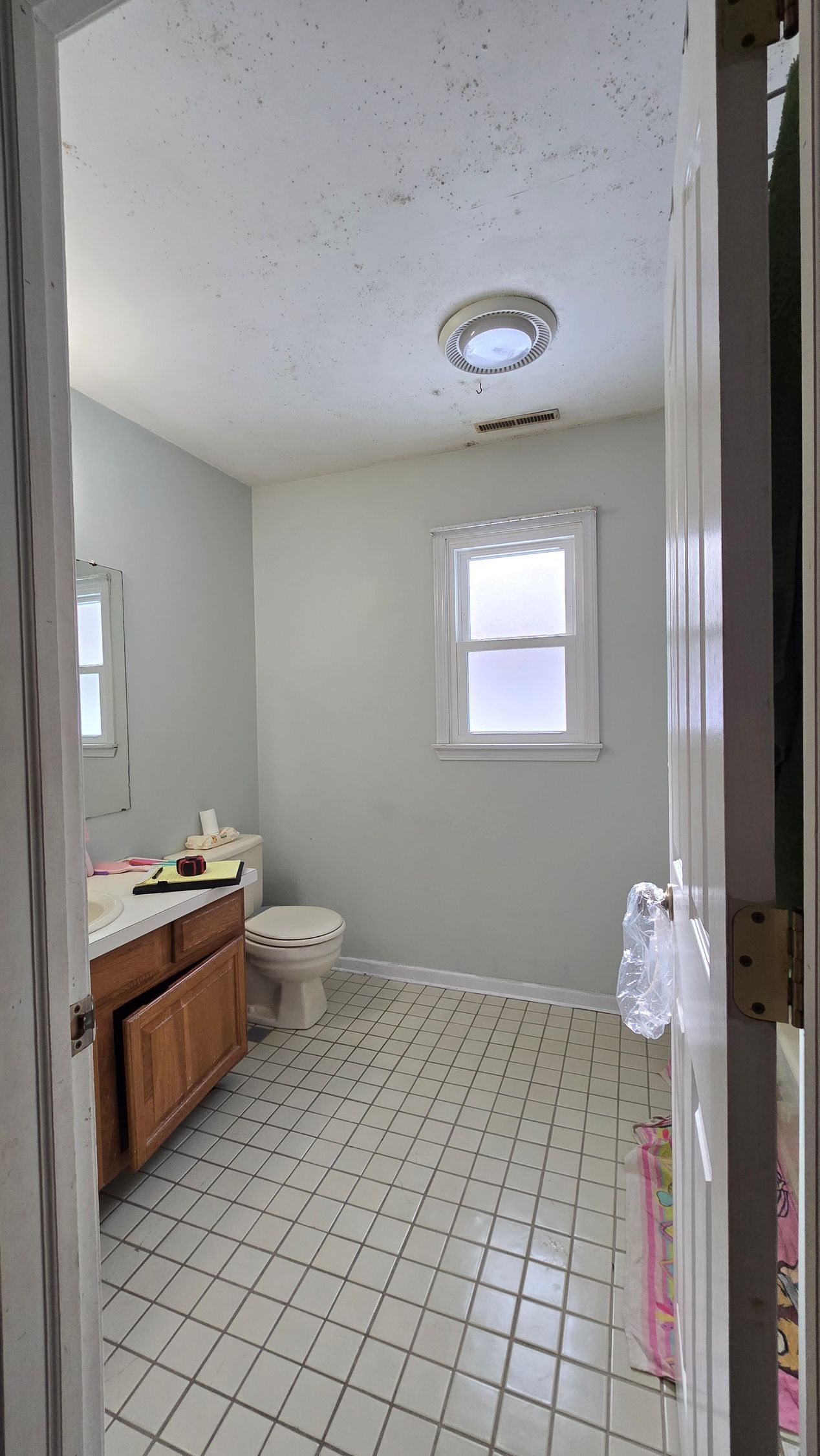 BEFORE Small bathroom with white walls, tiled floor, wood vanity, toilet, and a window with a lace valance.
