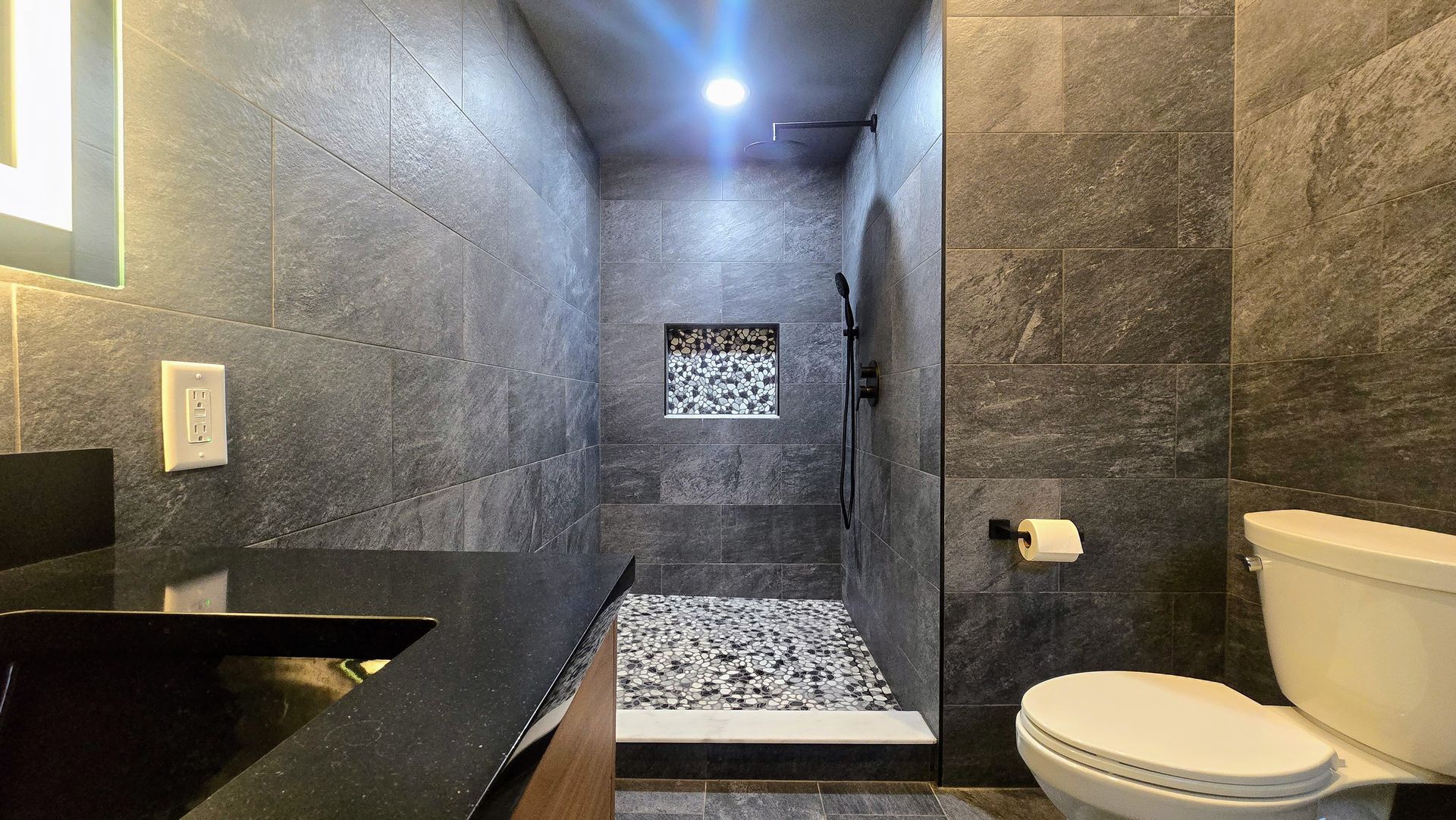 Gray tile shower with stone floor and built-in bench, next to a toilet.