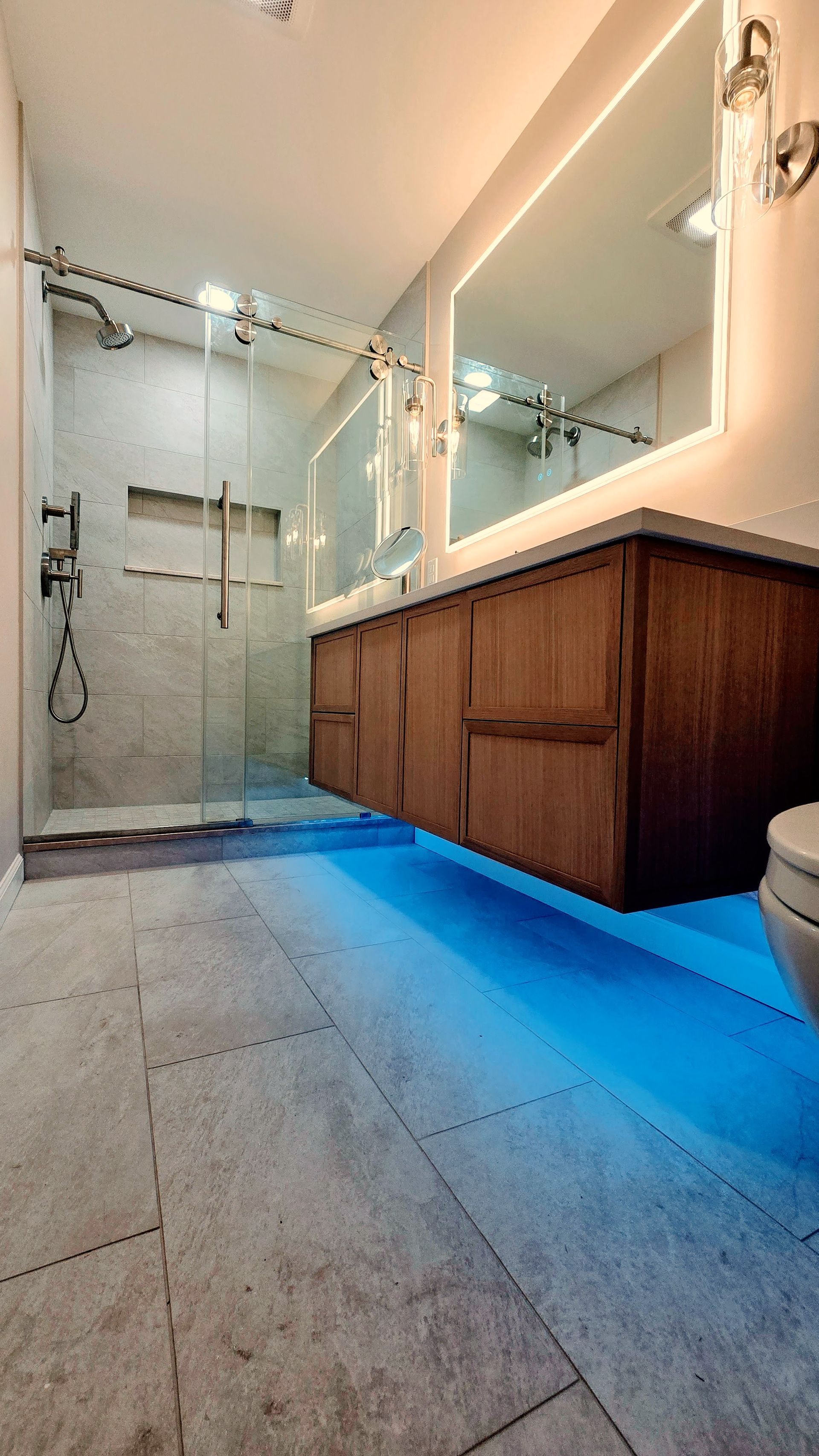 Modern bathroom in Simsbury, CT with wood vanity, blue LED floor lighting, glass-enclosed shower, and large wall mirror.