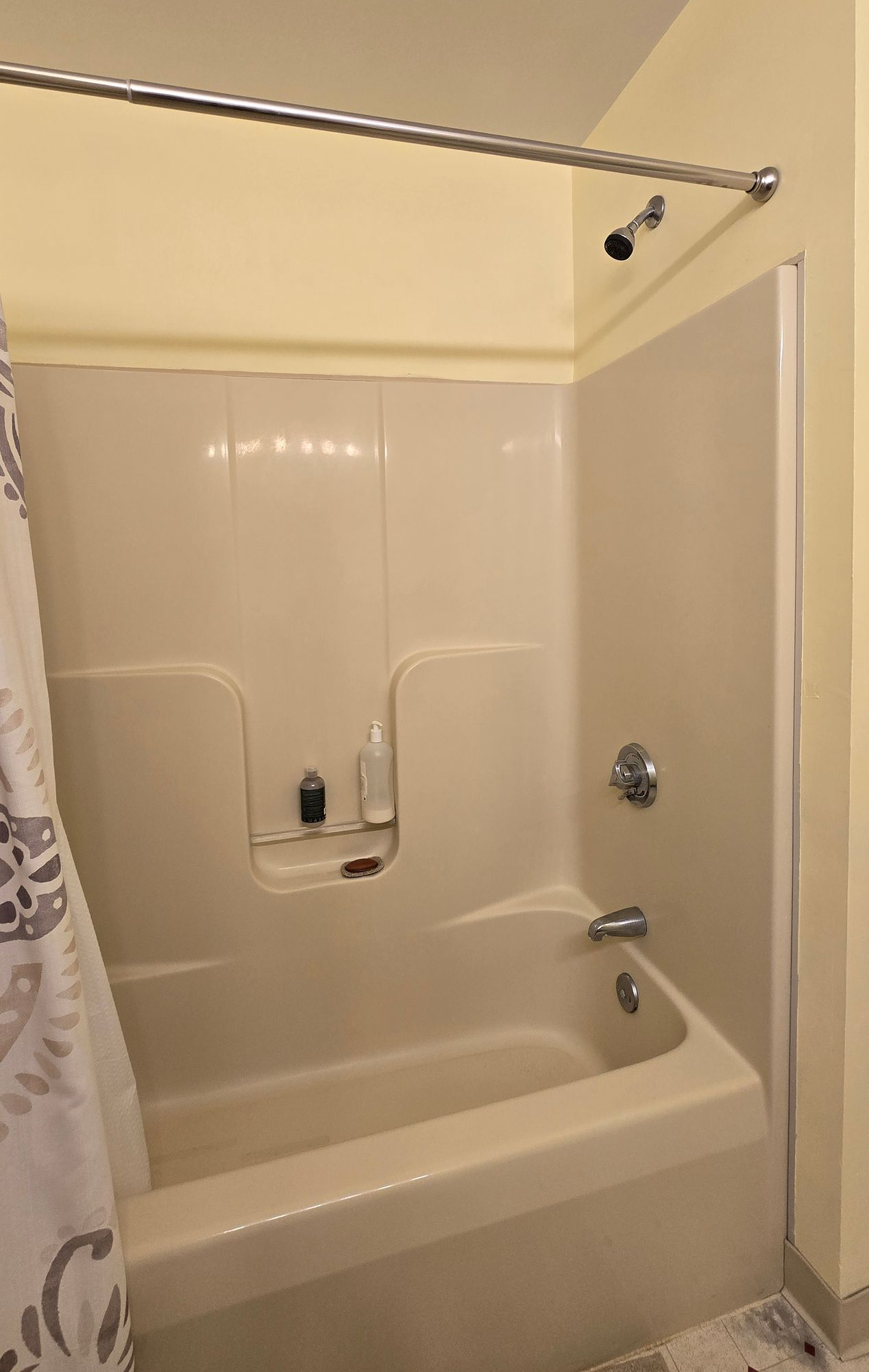 Bathtub shower with beige walls in Plainville, CT with curtain rod, grab bar, and built-in soap shelf in a small bathroom