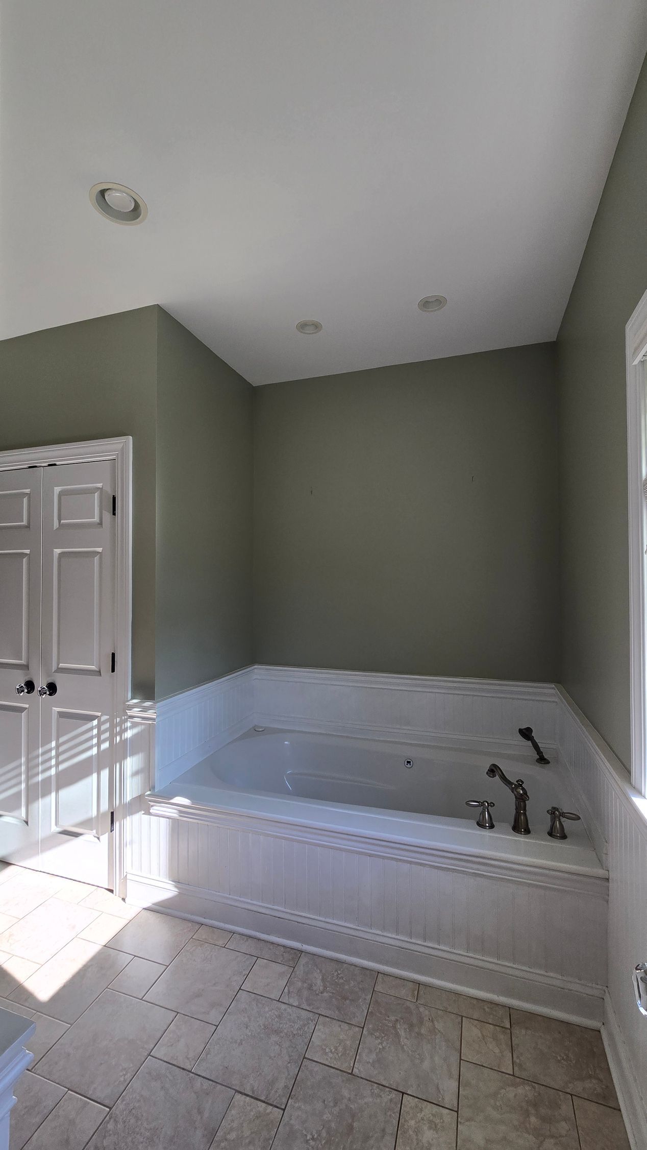 Empty bathroom with olive walls, white tile wainscoting, a tub, and an open door with sunlight. By First Choice Bathroom Remodeling done in Haddam, CT.