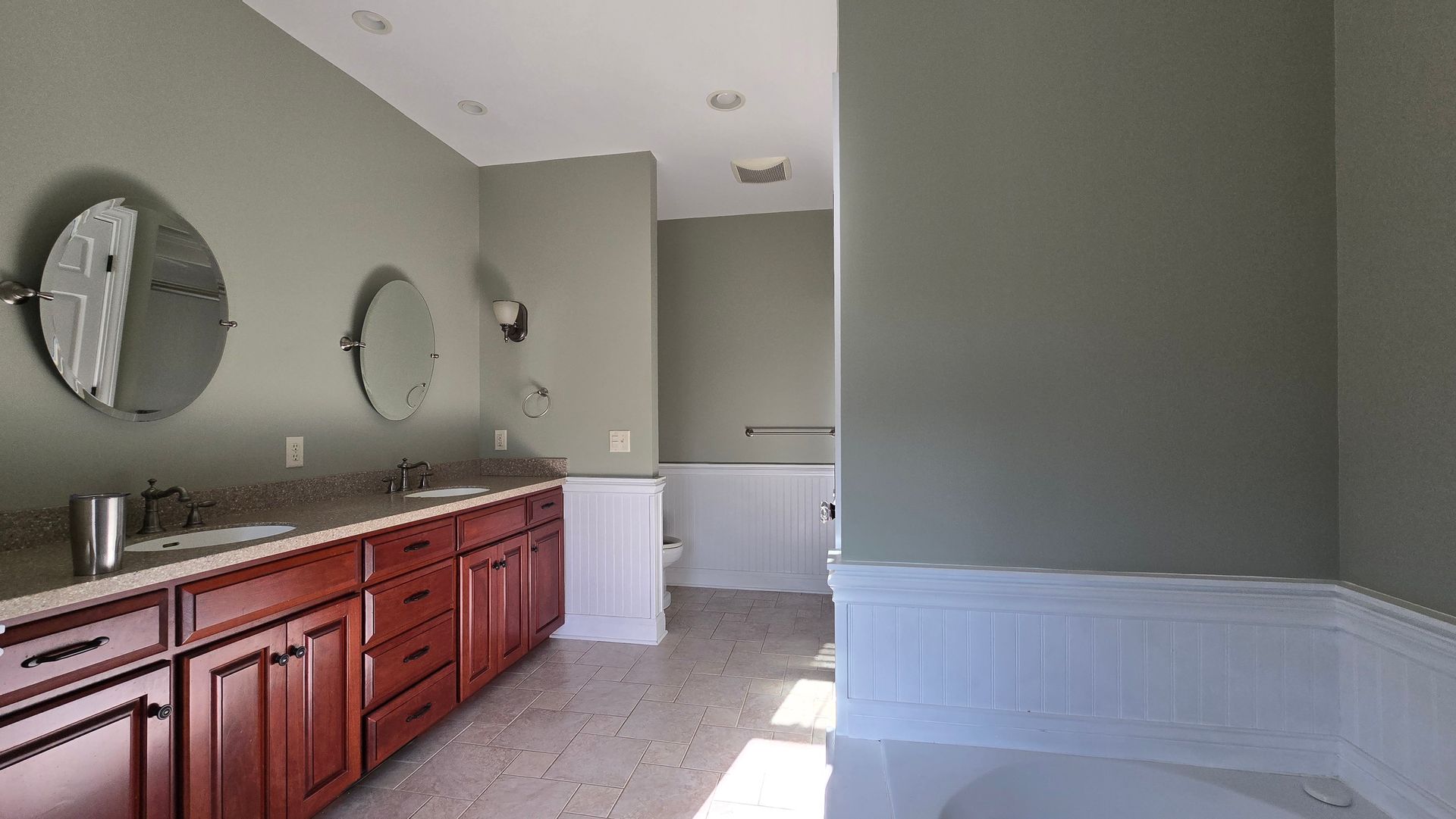 Bathroom with double sink vanity, round mirrors, gray walls, and white wainscoting. By First Choice Bathroom Remodeling done in Haddam, CT.