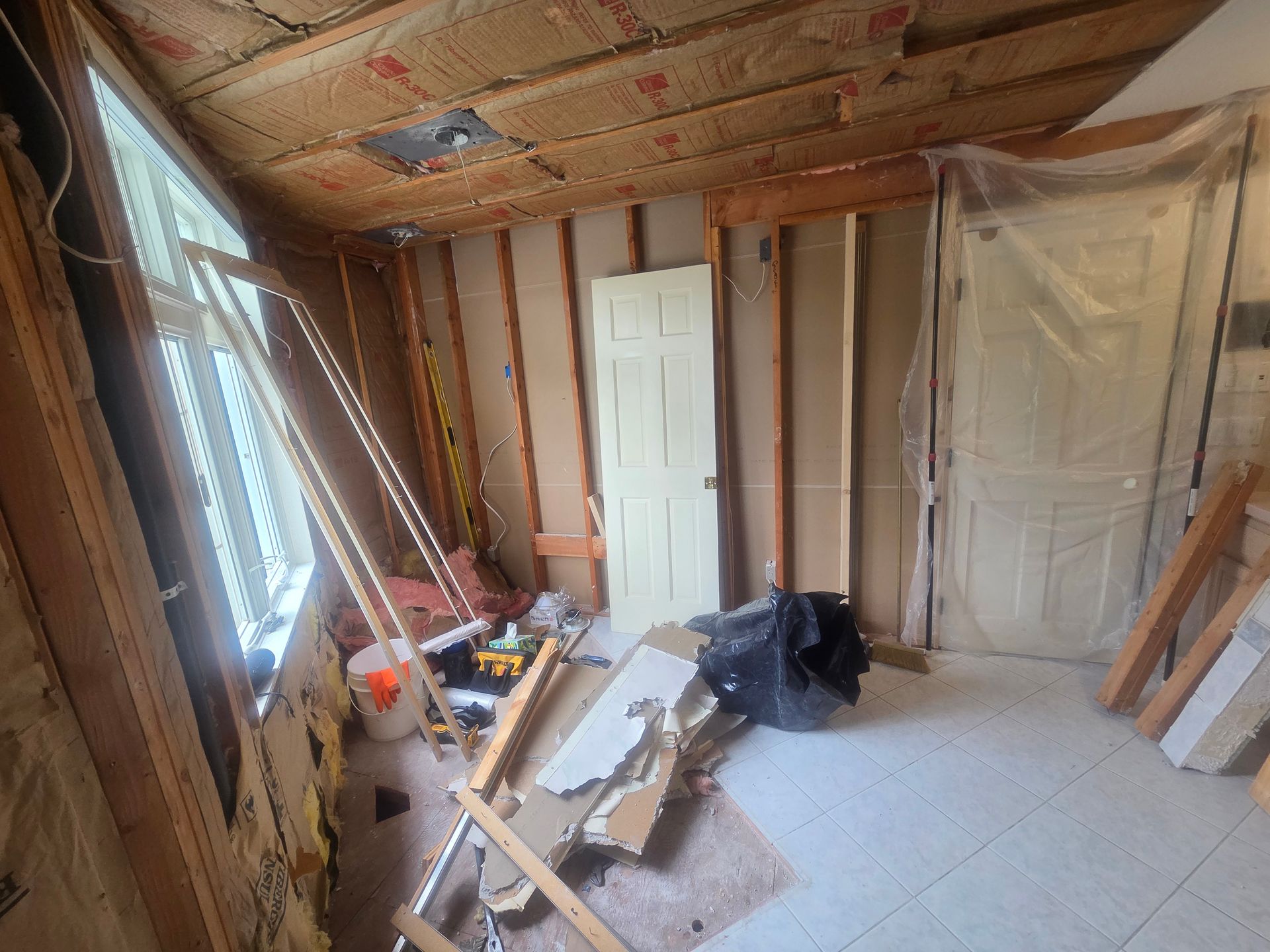 Renovation in progress inside a bare room with exposed studs, white doors, tools, and debris on the floor