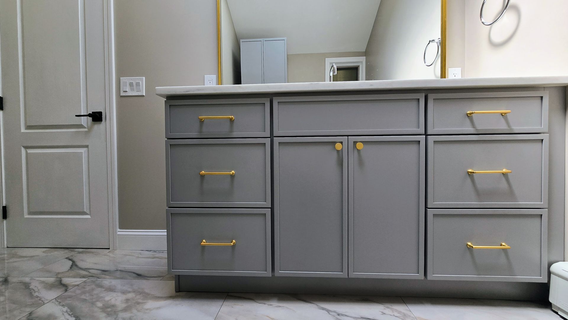 Gray bathroom vanity with gold handles, double sinks, and a large mirror above a marble-pattern floor.