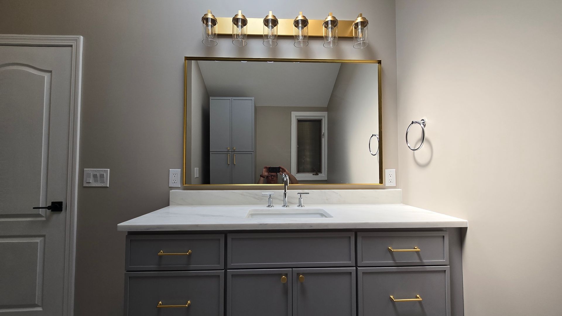 Modern bathroom vanity with white countertop, gray cabinets, gold-framed mirror, and four-light fixture above.