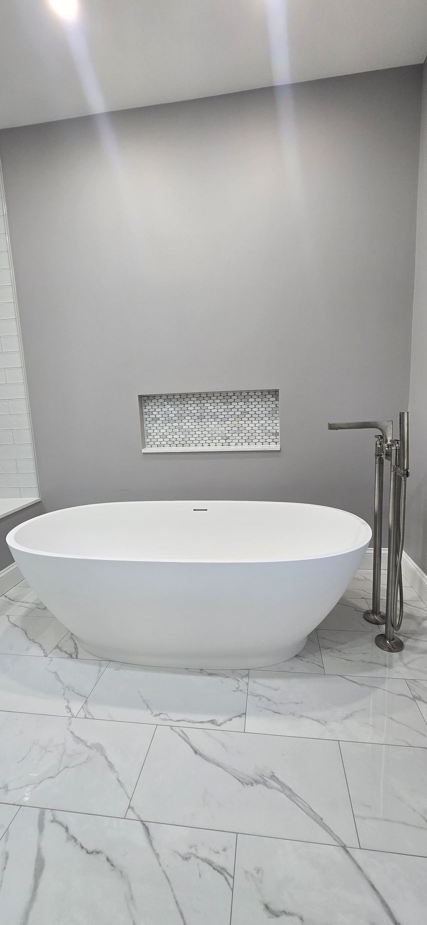 Modern bathroom with a white oval tub, gray walls, marble floor, and chrome fixtures. By First Choice Bathroom Remodeling done in Haddam, CT.