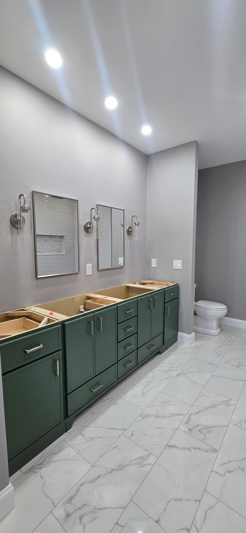 Modern bathroom with green vanities, wall mirrors, bright lights, and a white toilet on marble tile floor. By First Choice Bathroom Remodeling done in Haddam, CT.