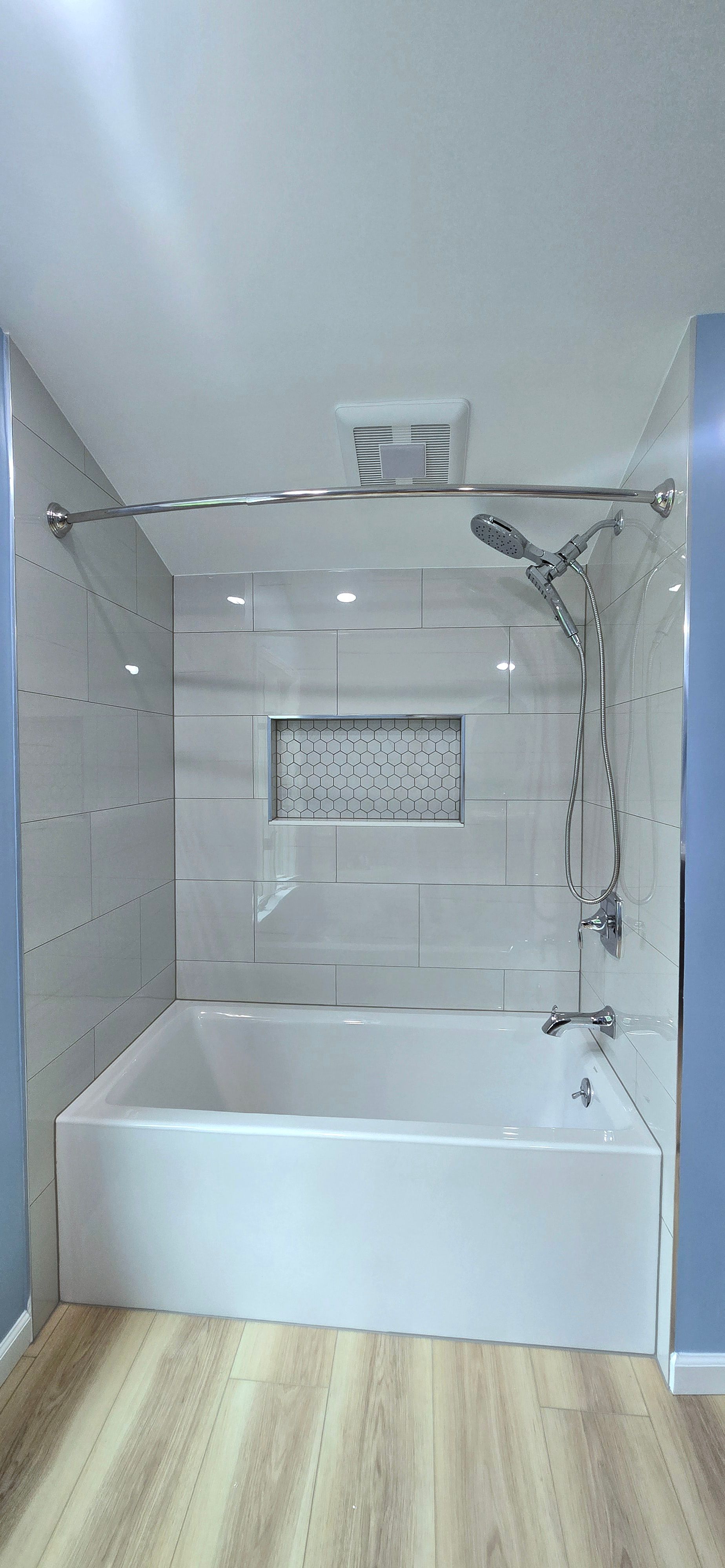 White tiled bathtub shower alcove with chrome fixtures, glass shelves, and a frosted window above.