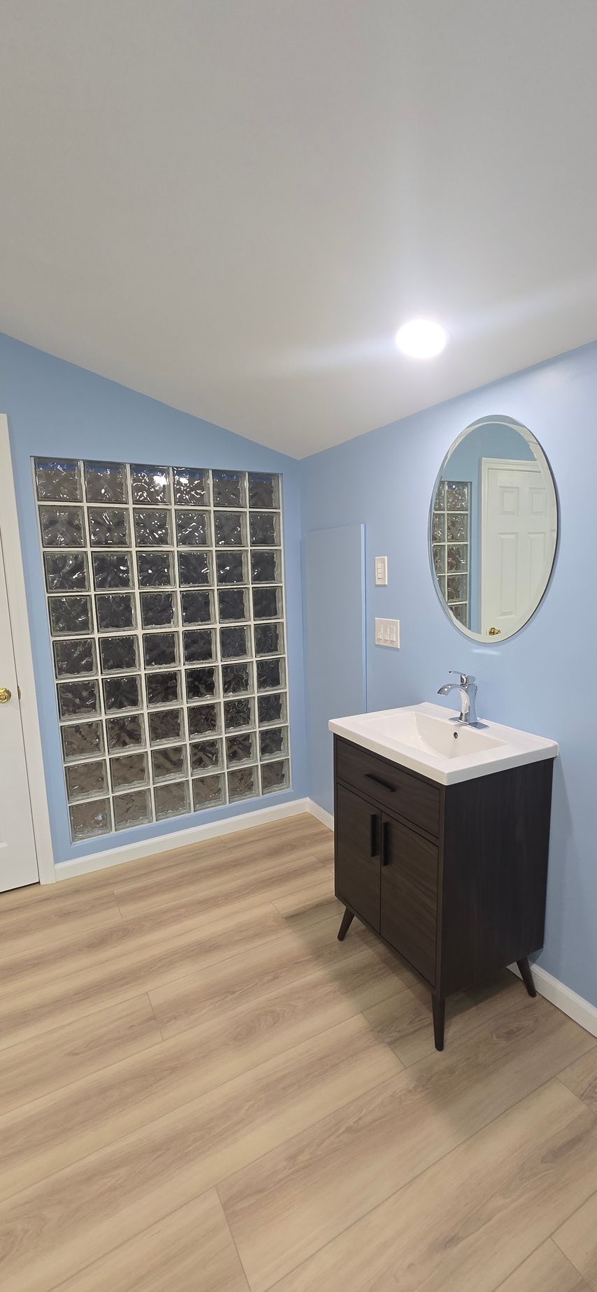 Blue-tiled bathroom with glass-block shower wall, oval mirror, and dark vanity on light wood floor.