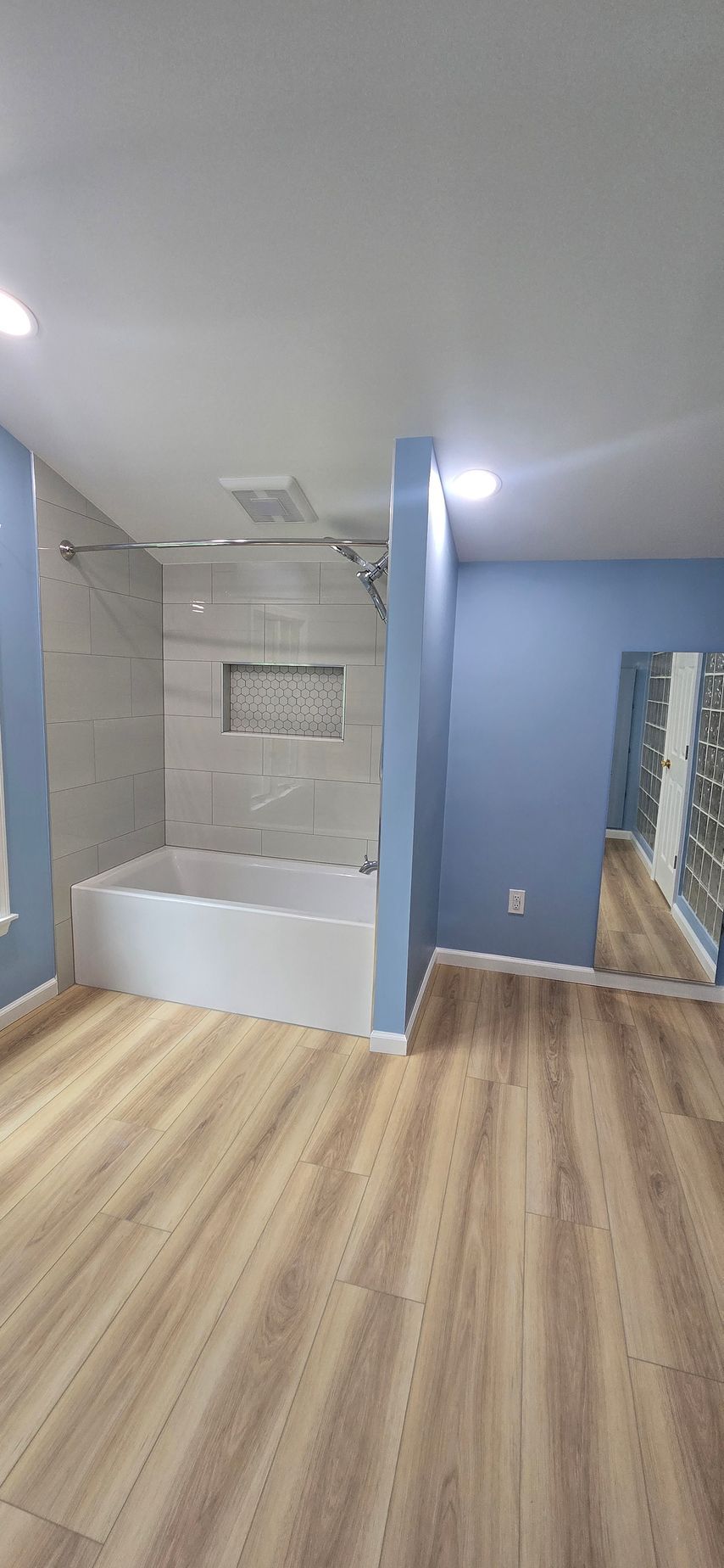 Bathroom with white tub and tiled shower beside blue walls and wood-look flooring