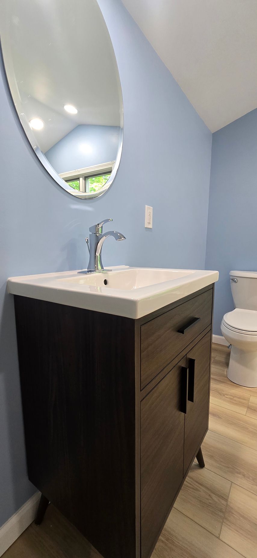 Modern bathroom vanity with oval mirror, white sink, blue walls, and a toilet in the corner