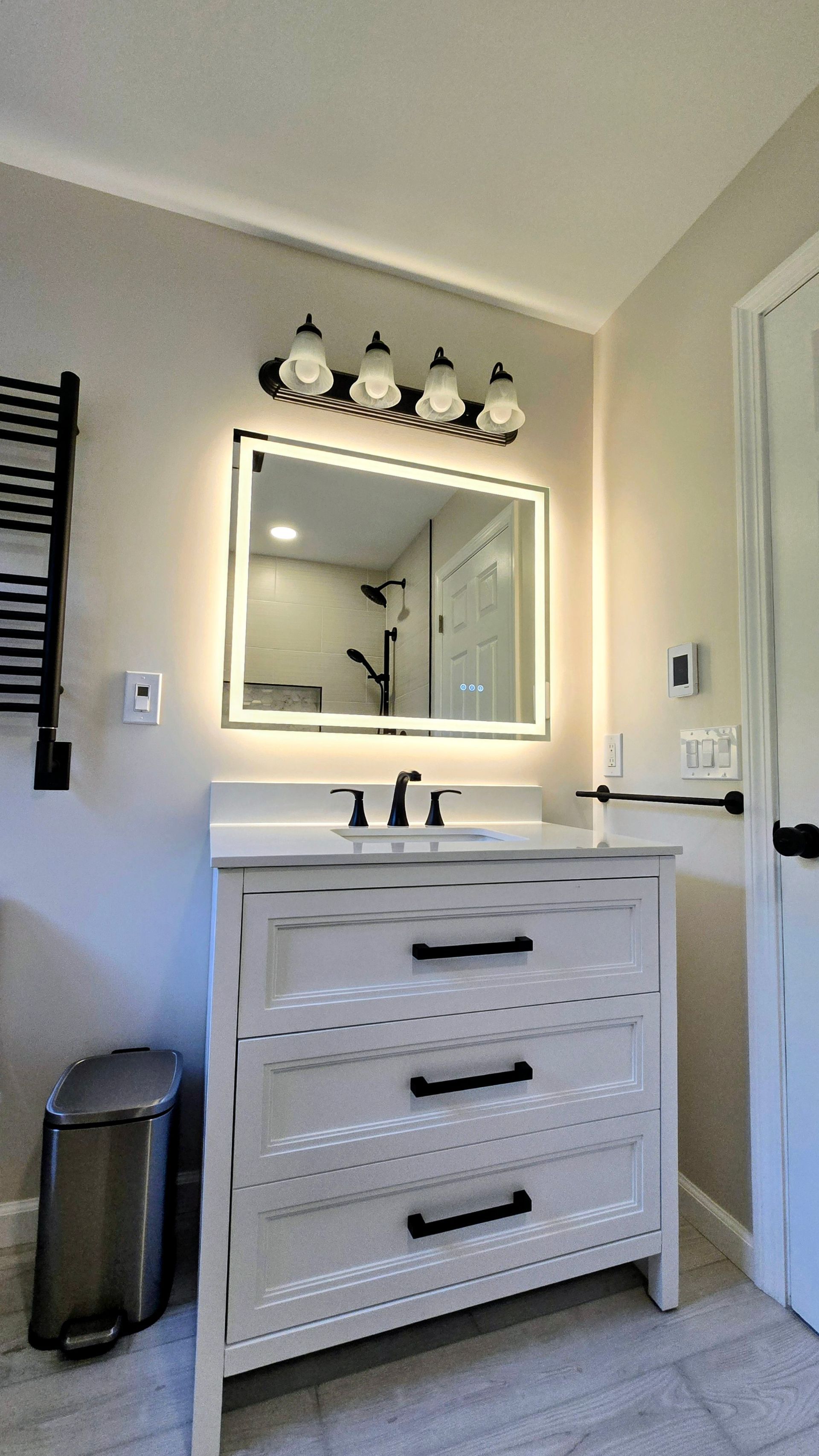 White bathroom vanity with lit mirror, black fixtures, and trash can beside a closed door.