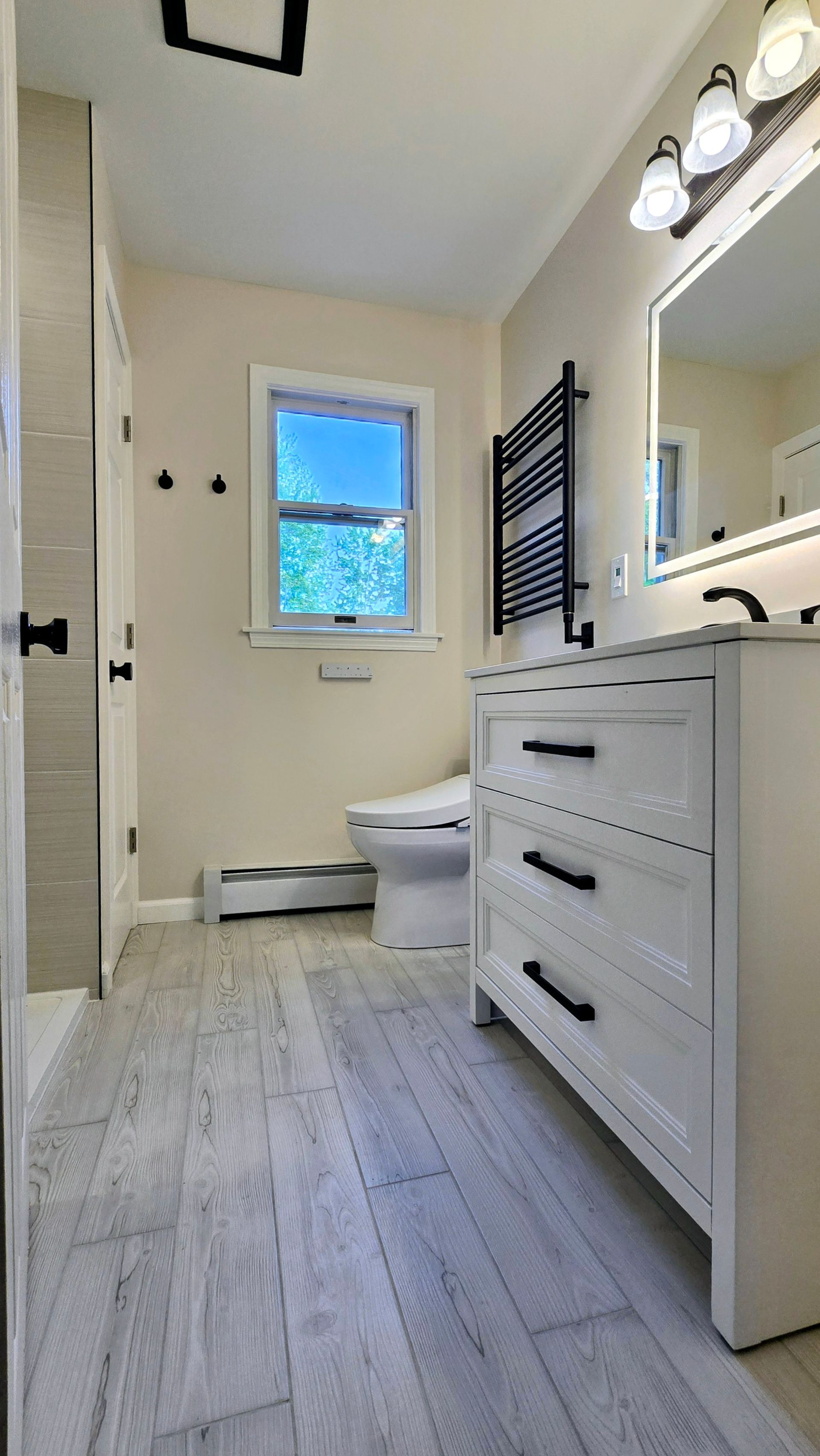 Bright bathroom with white vanity, mirror, black towel rack, and window above the toilet.