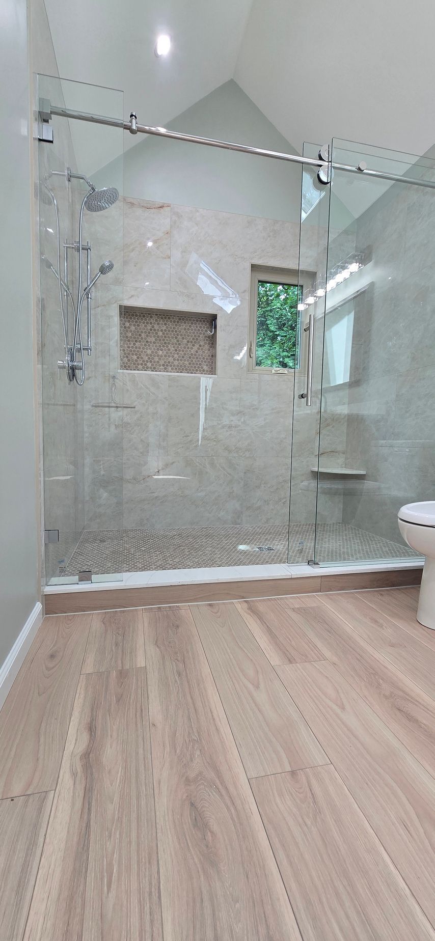 Small empty room with glass shower stall, light wood floor, and bright window.