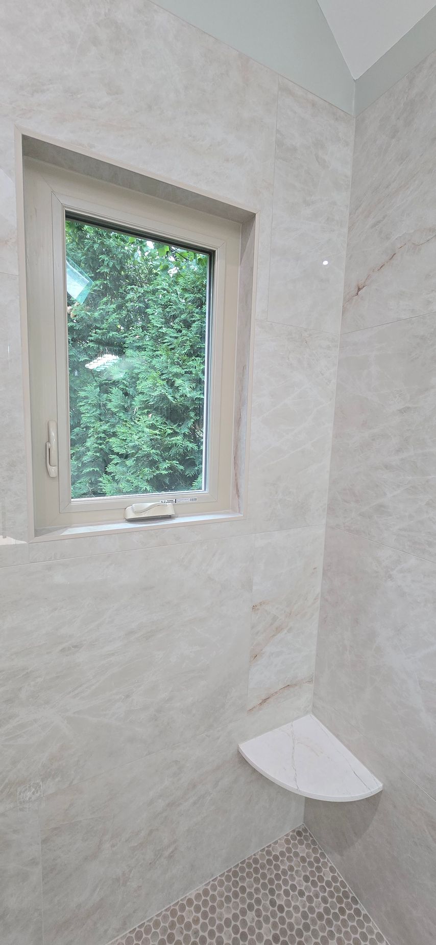 Small tiled bathroom with a window and a corner shower seat, lit by daylight.
