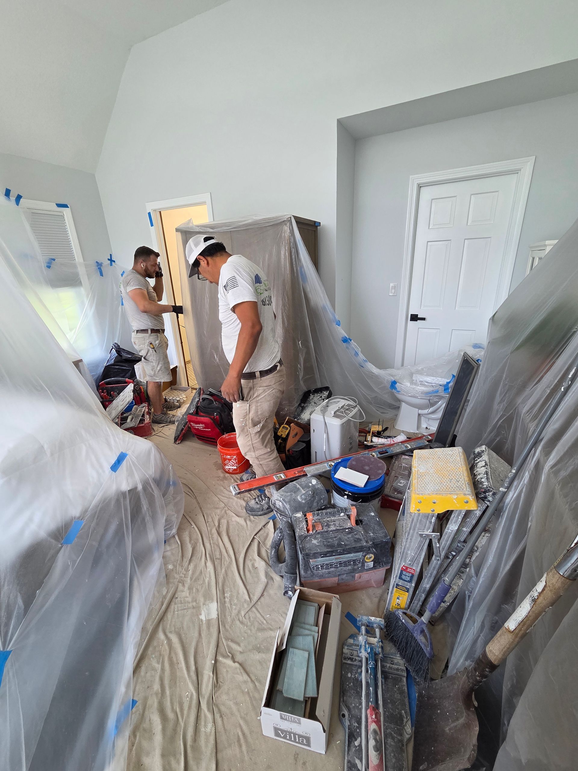 Two workers in a plastic-covered room amid renovation tools and debris.