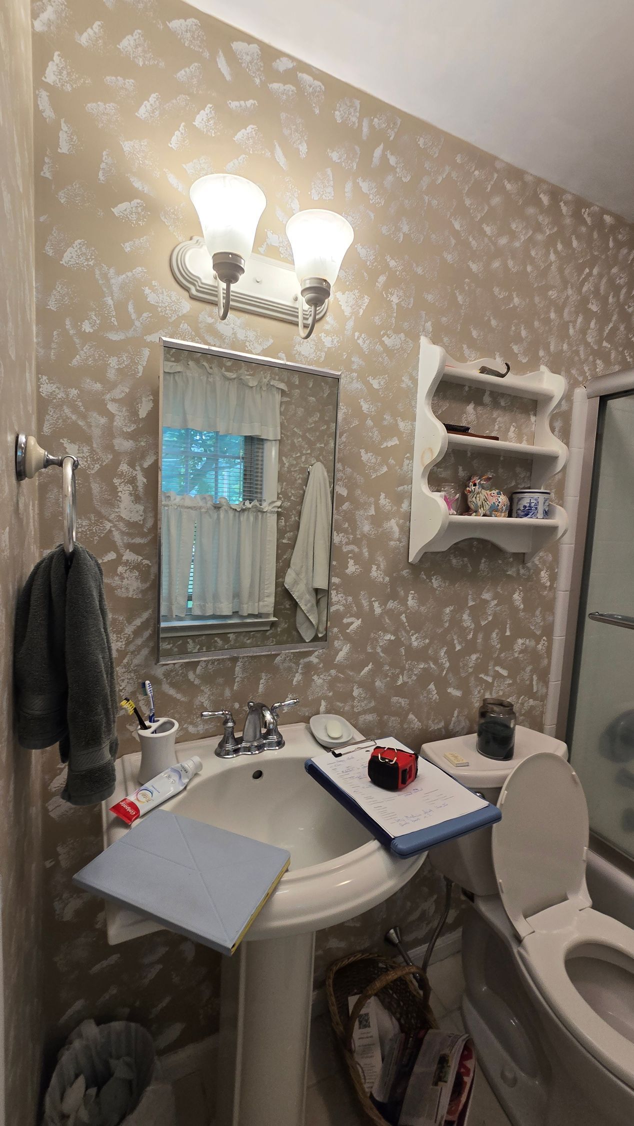 Small bathroom with a sink, mirror, wall sconce, toilet, and shelves in beige wallpaper.