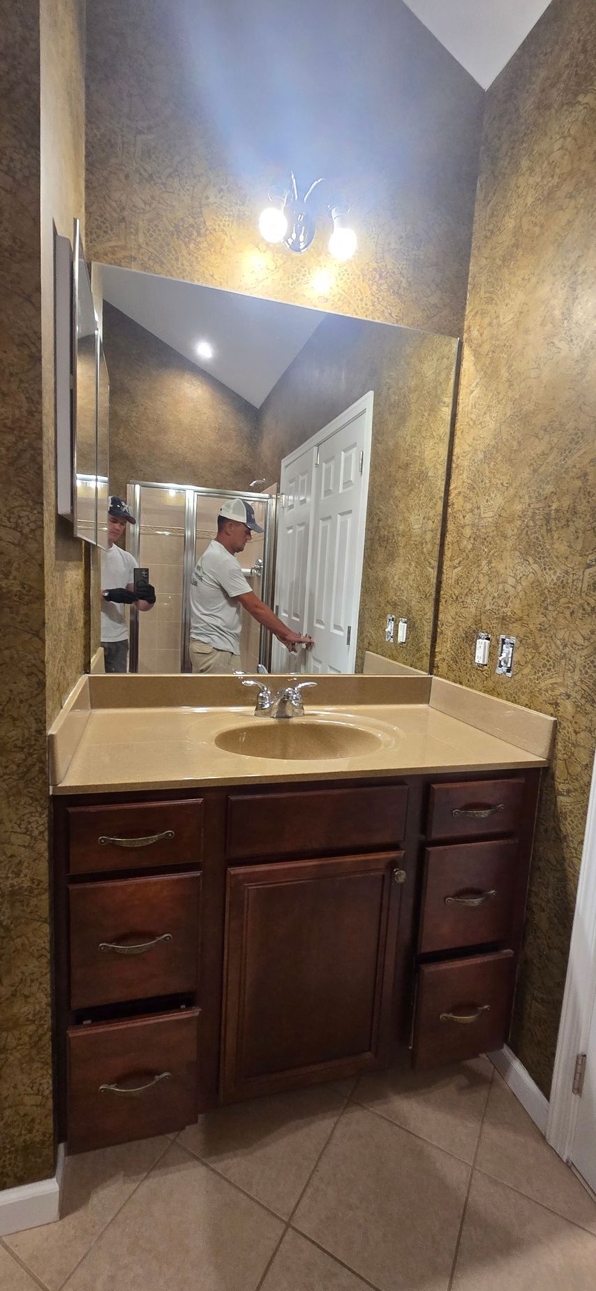 Narrow bathroom with granite vanity, mirror, and wall lights; a person stands reflected near the sink.