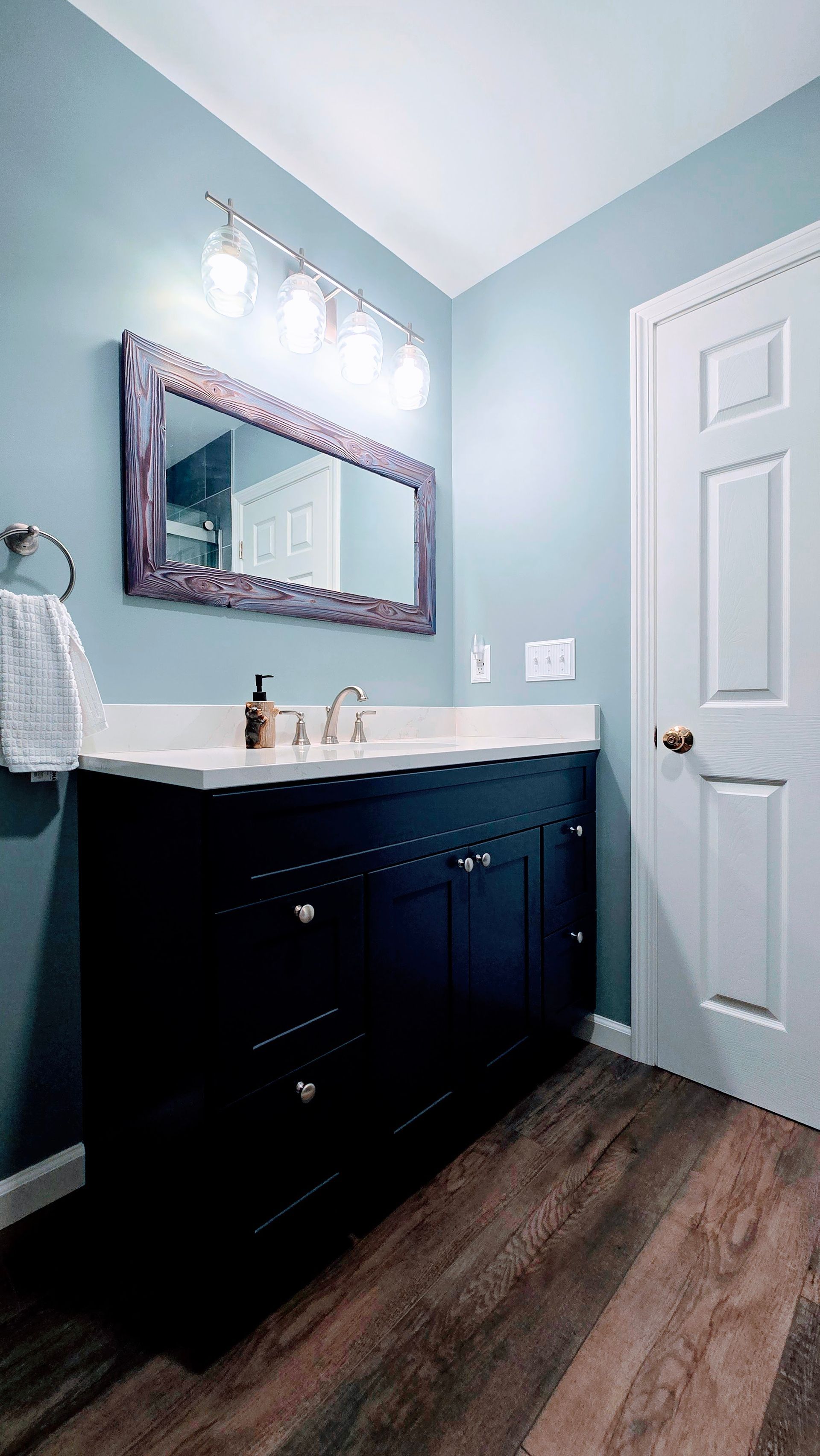 Bright bathroom with light blue walls, dark vanity, white sink, framed mirror, and wood-look flooring.