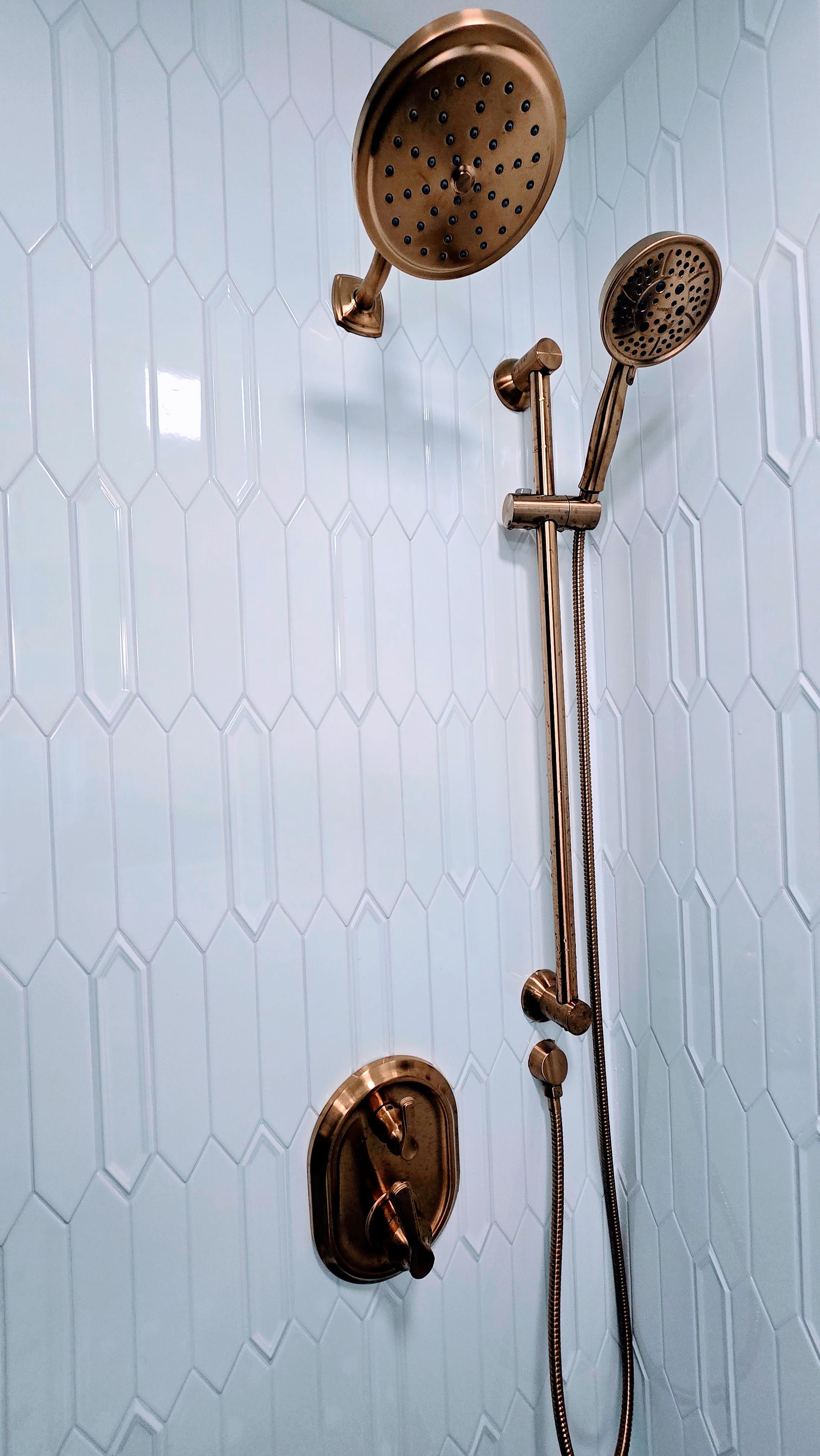 Brass shower fixtures on a white tiled wall with a hand shower and rain showerhead. Done by First Choice Bathroom Remodeling done in Manchester, CT.