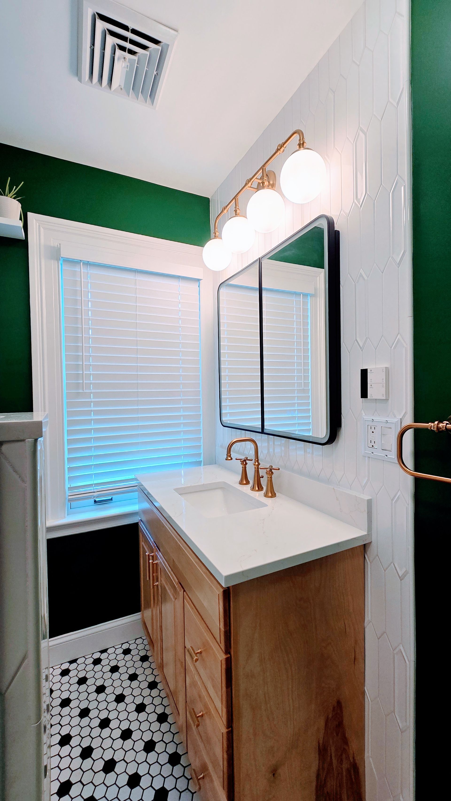 Small bathroom with wood vanity, round mirror, white sink, and window with blinds and green accent wall. Done by First Choice Bathroom Remodeling done in Manchester, CT.