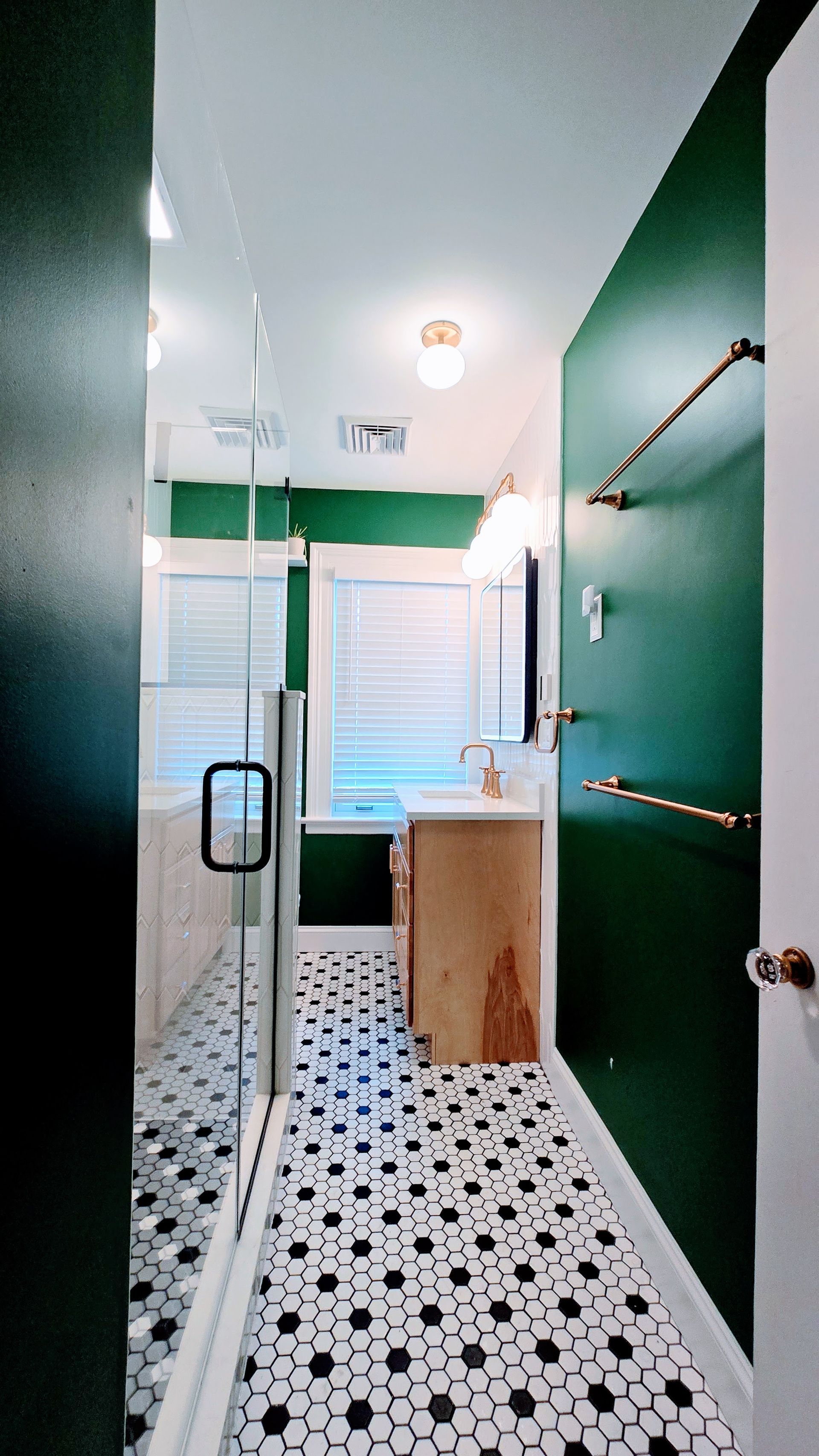 Narrow bathroom with green walls, black-and-white tile floor, glass shower, and a window at the far end