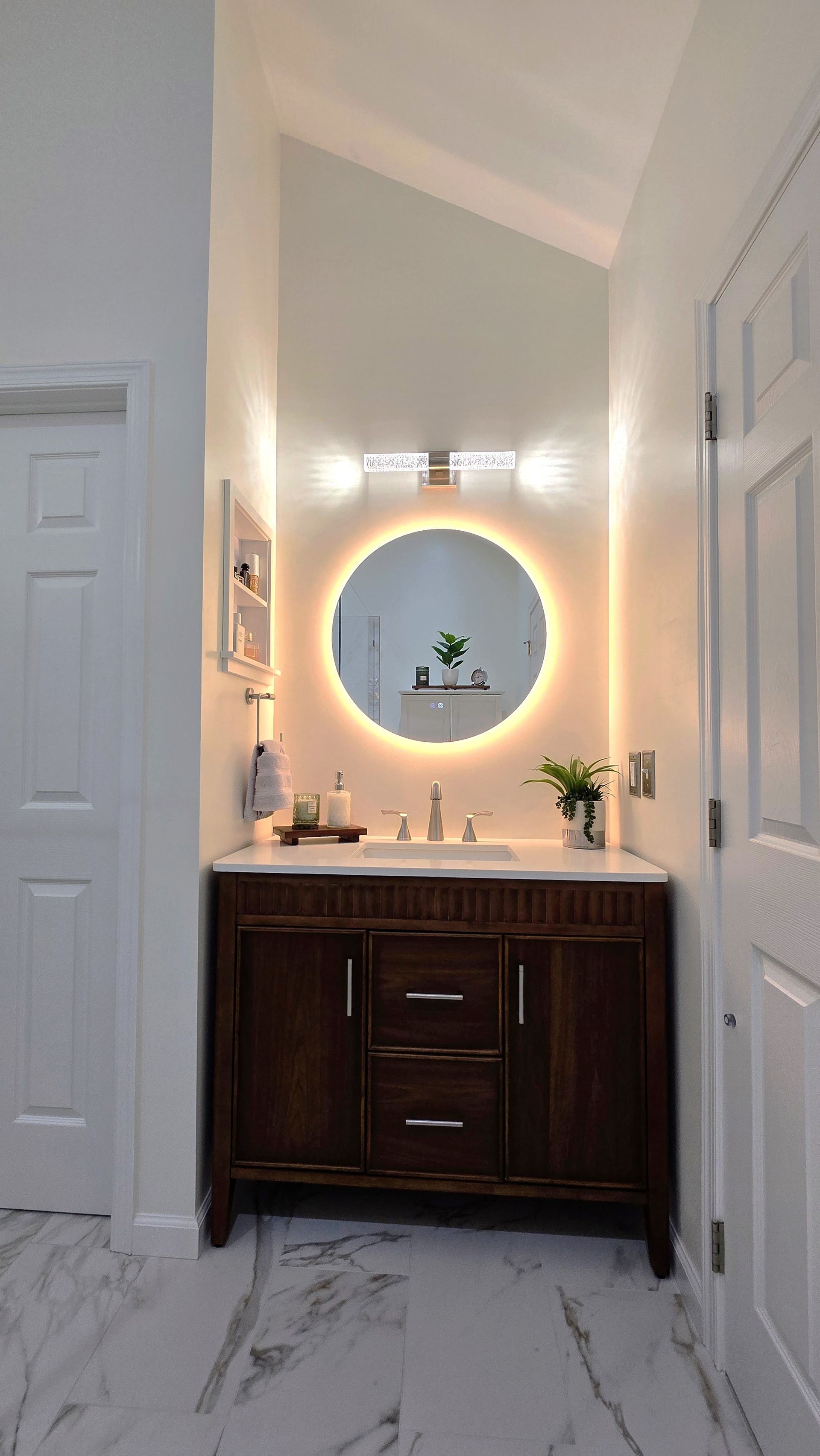 Modern bathroom remodeling completed by First Choice Bathroom Remodeling vanity with round backlit mirror, dark wood cabinet, marble floor, and white walls