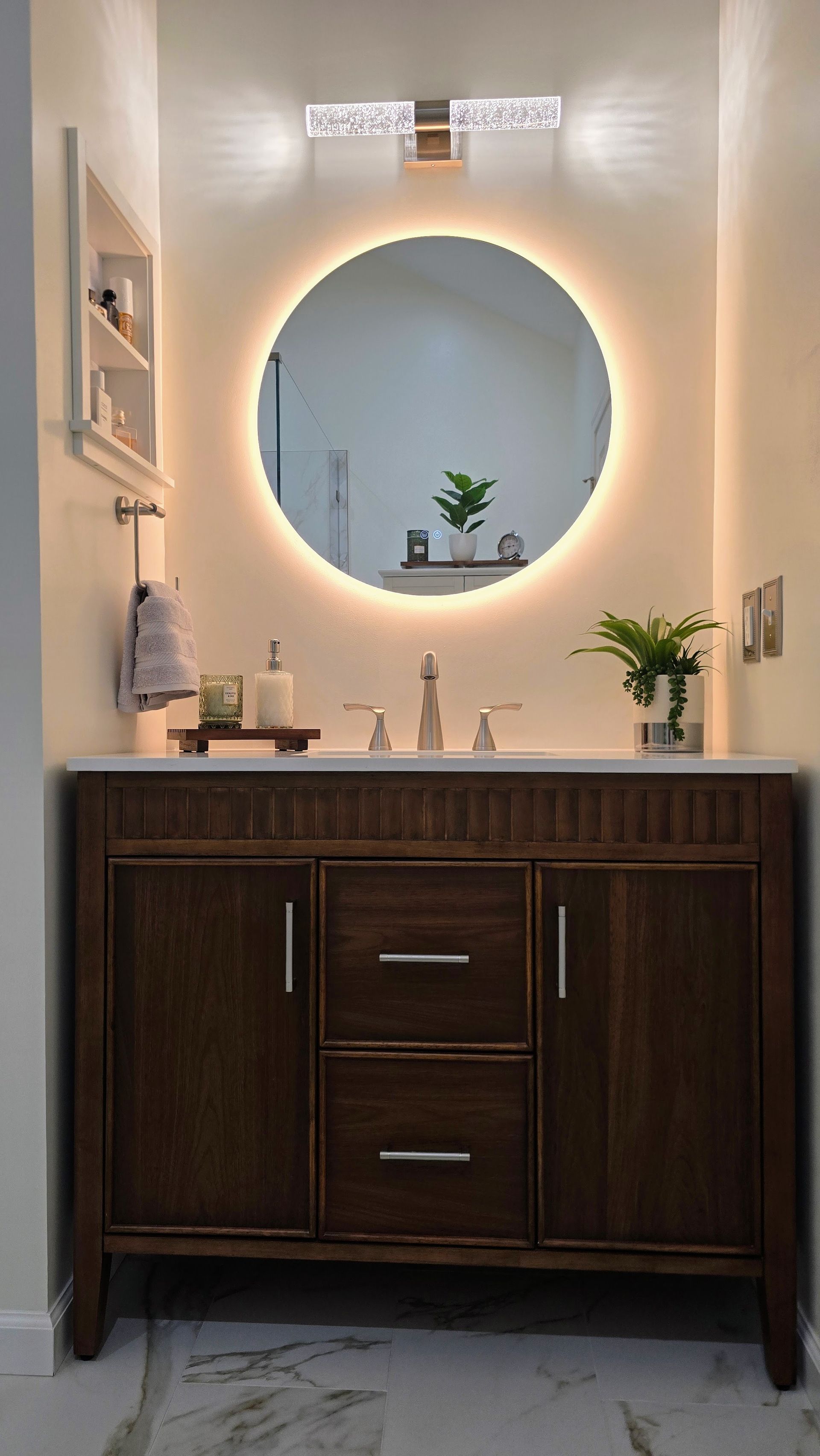 Bathroom vanity with wood cabinet, round backlit mirror, and plants on a narrow wall niche. By First Choice Bathroom Remodeling done in Prospect, CT.