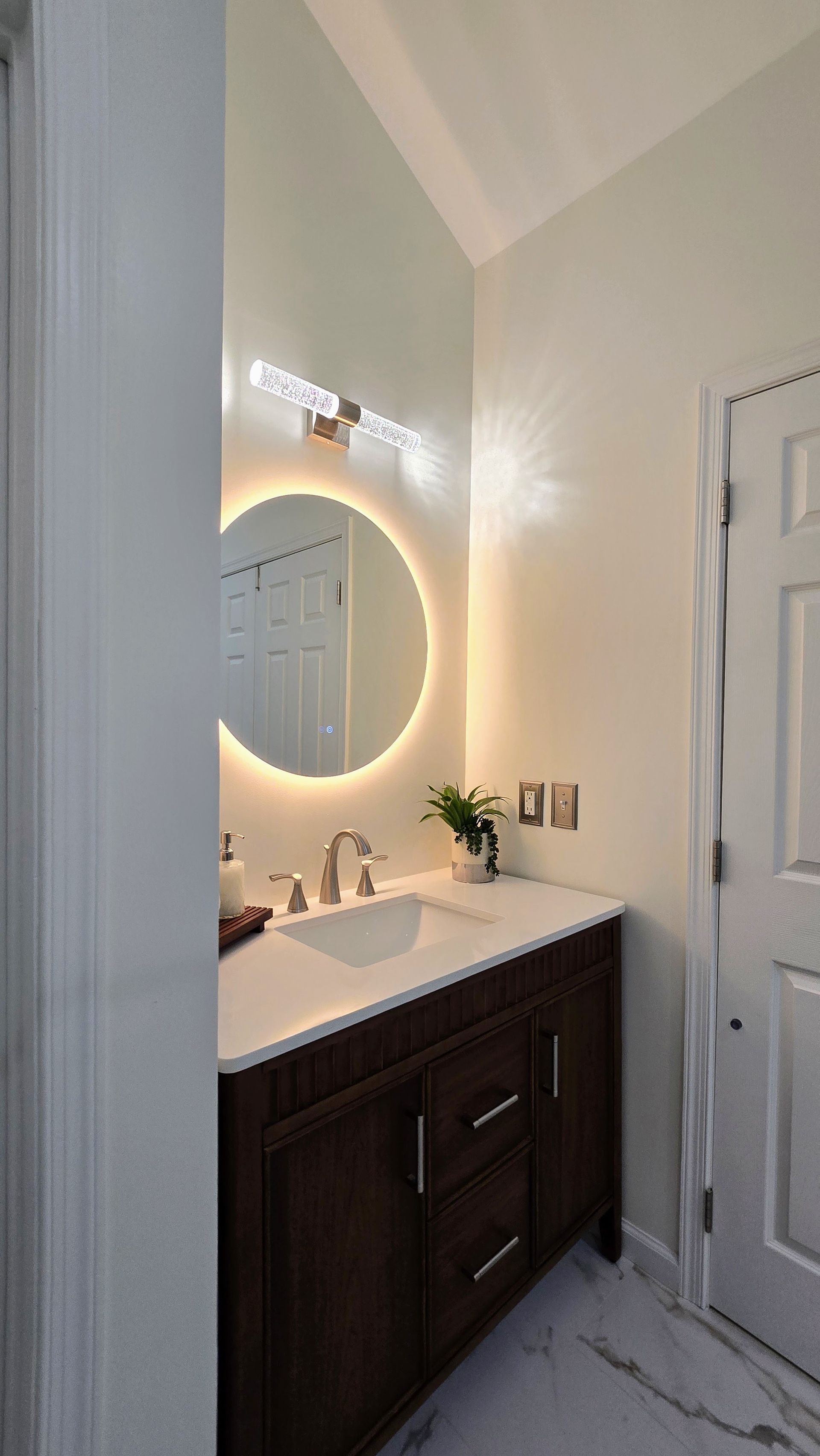 Small bathroom vanity with lit round mirror, white sink, dark cabinet, and white door on marble-look floor. By First Choice Bathroom Remodeling done in Prospect, CT.
