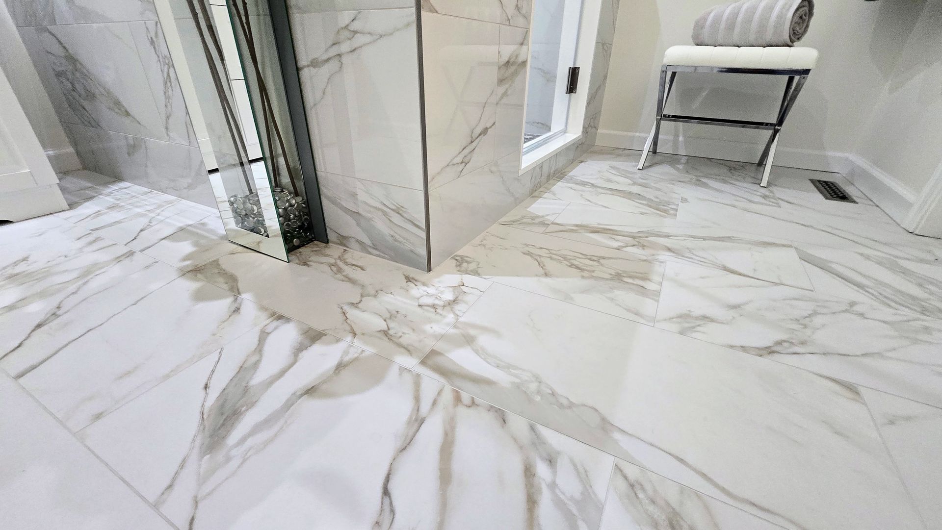 Luxurious white marble bathroom with a glass shower, modern chair, and gold-veined floor tiles. By First Choice Bathroom Remodeling done in Prospect, CT.
