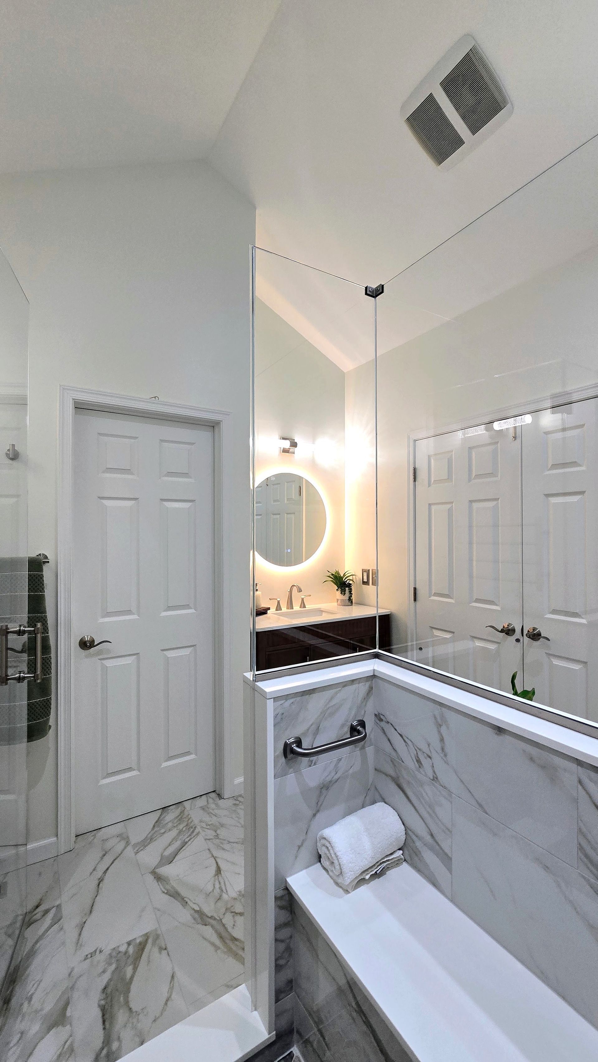 Bright white bathroom completed by First Choice Bathroom Remodeling with marble walls, double doors, round mirror, and a bathtub in the foreground.