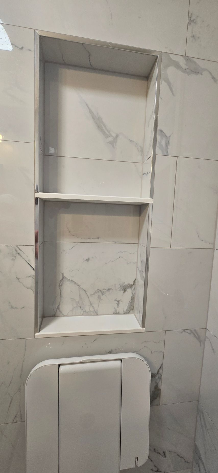 Built-in wall niche with white marble-look tiles above a toilet paper holder in a tiled bathroom. By First Choice Bathroom Remodeling done in Cheshire, CT.