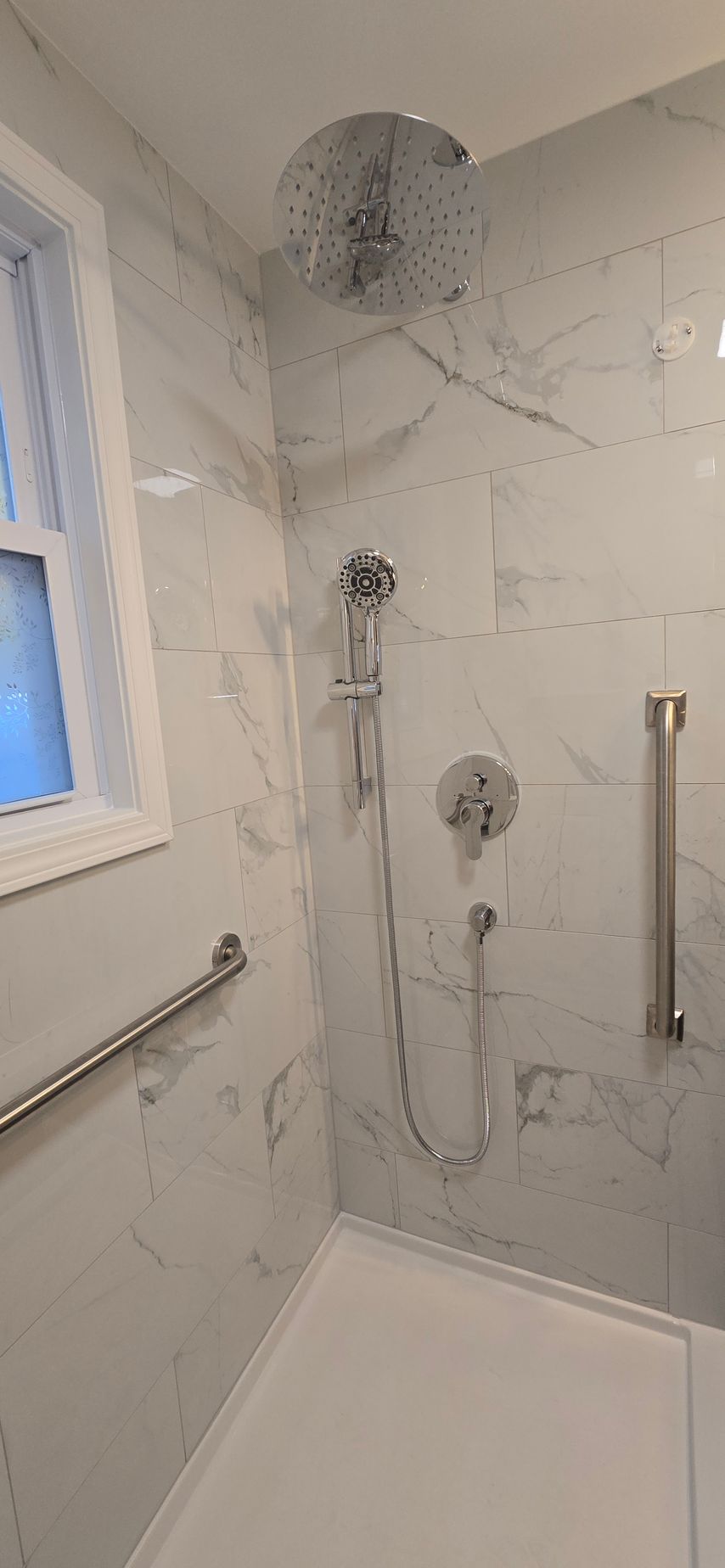 White tiled shower stall with rainfall showerhead, handheld sprayer, grab bars, and built-in bench seat. By First Choice Bathroom Remodeling done in Cheshire, CT.