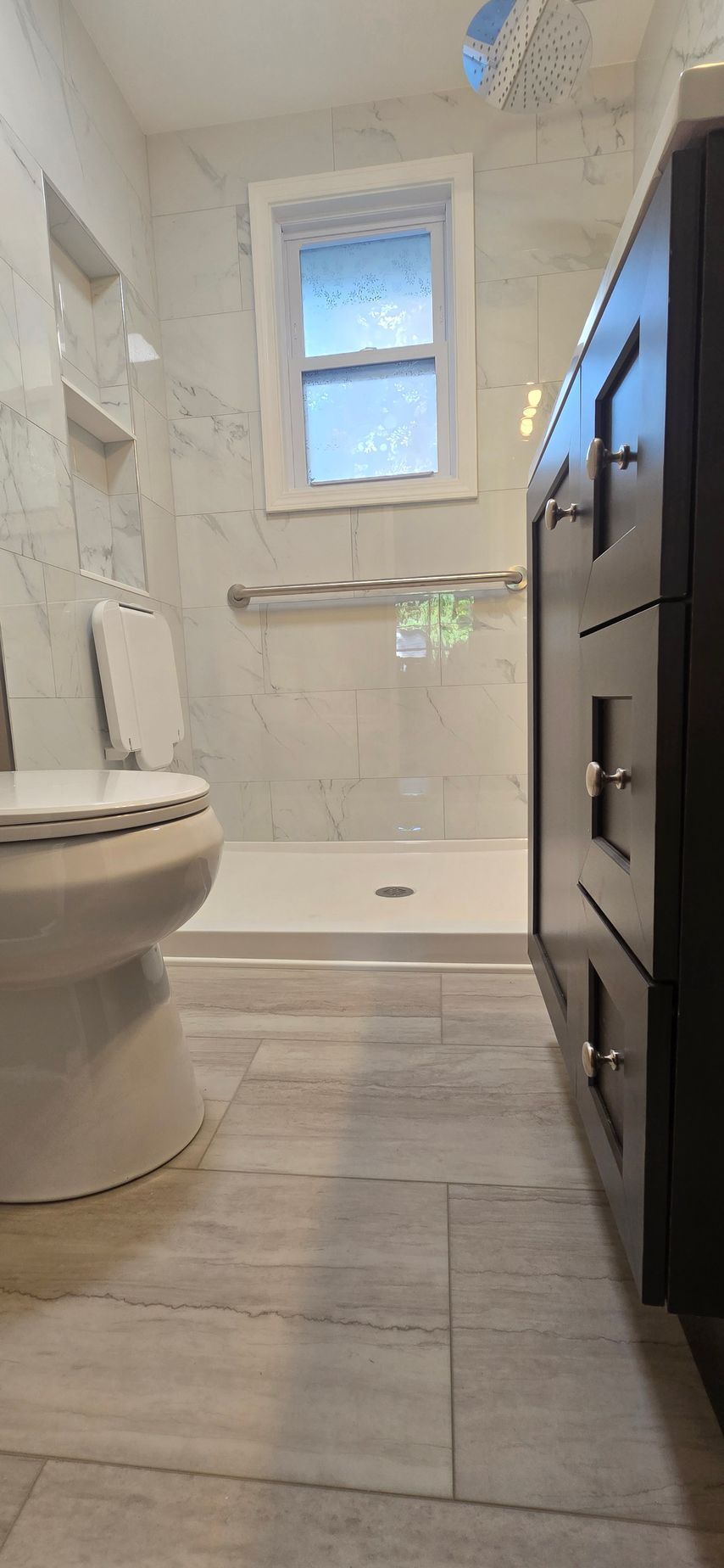 Narrow modern bathroom with bathtub, toilet, and dark vanity beside a small window. By First Choice Bathroom Remodeling done in Cheshire, CT.